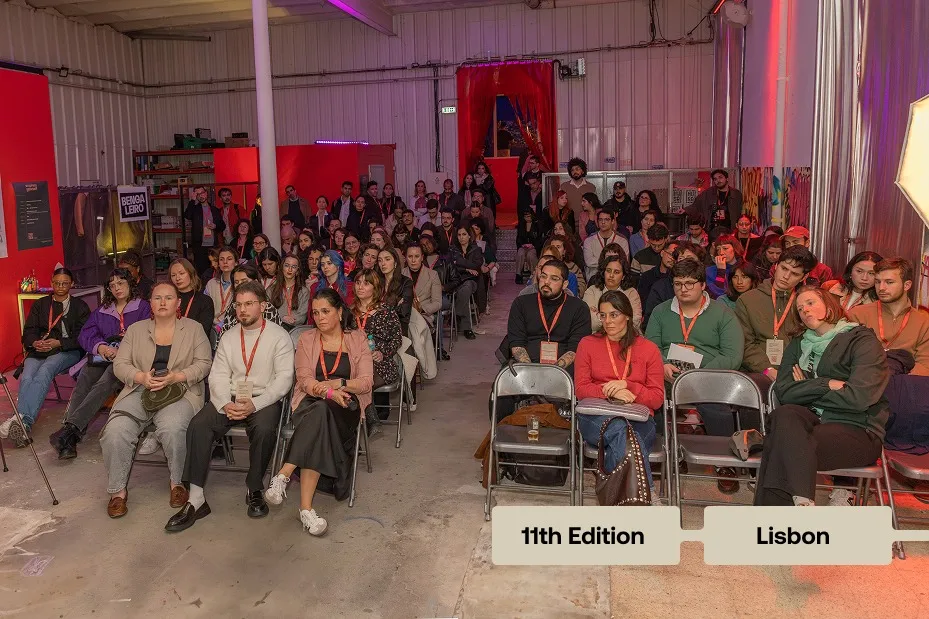 Large group of people seated indoors facing forward, attending an event in a warehouse-like space.