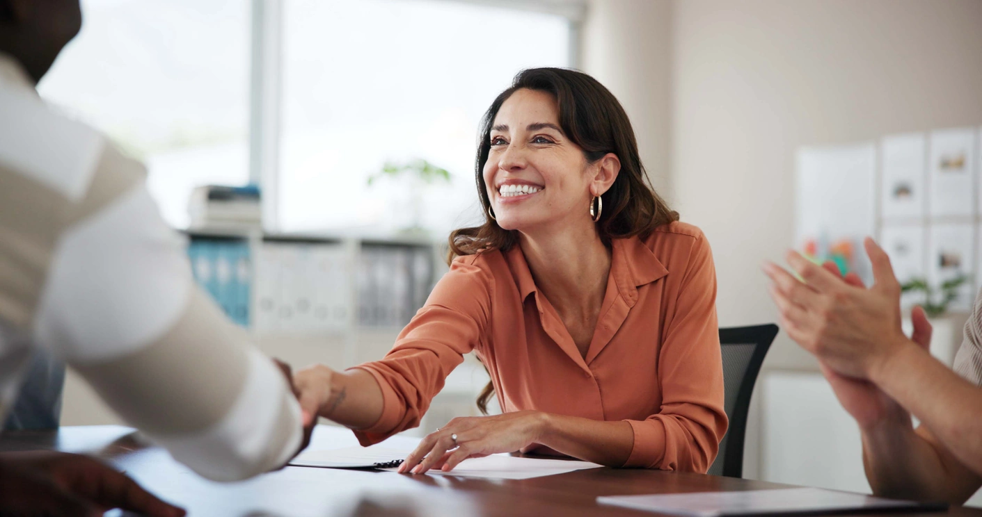 Smiling woman in an orange blouse shaking hands with a person across a table in an office setting while another person claps.