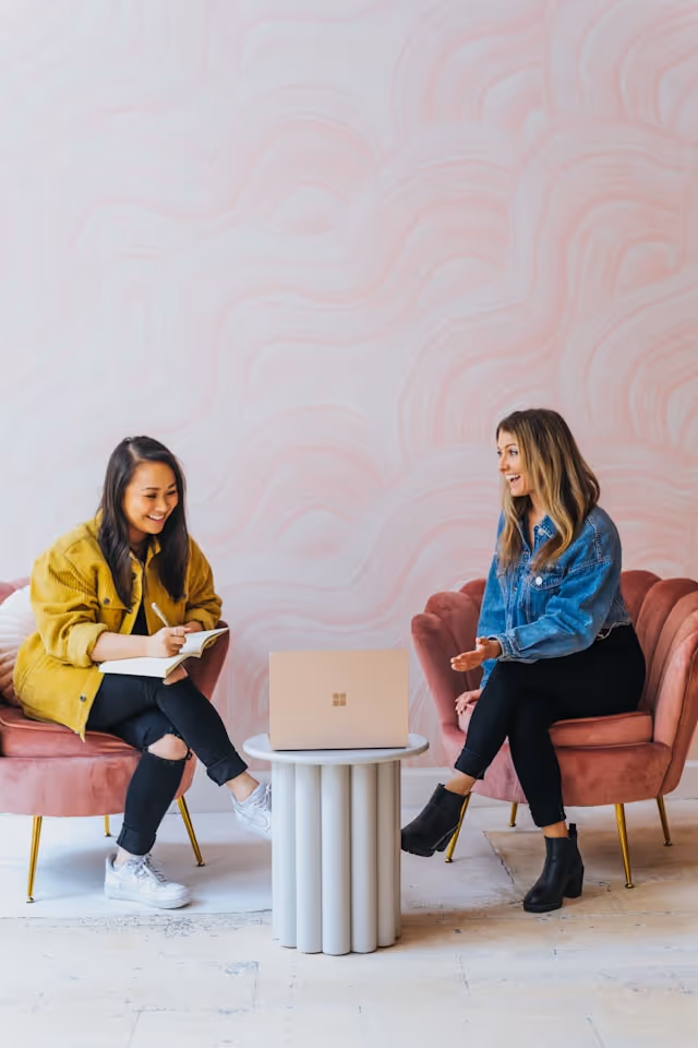 Two women sitting on pink velvet chairs conversing over a laptop on a small round table in a modern room.