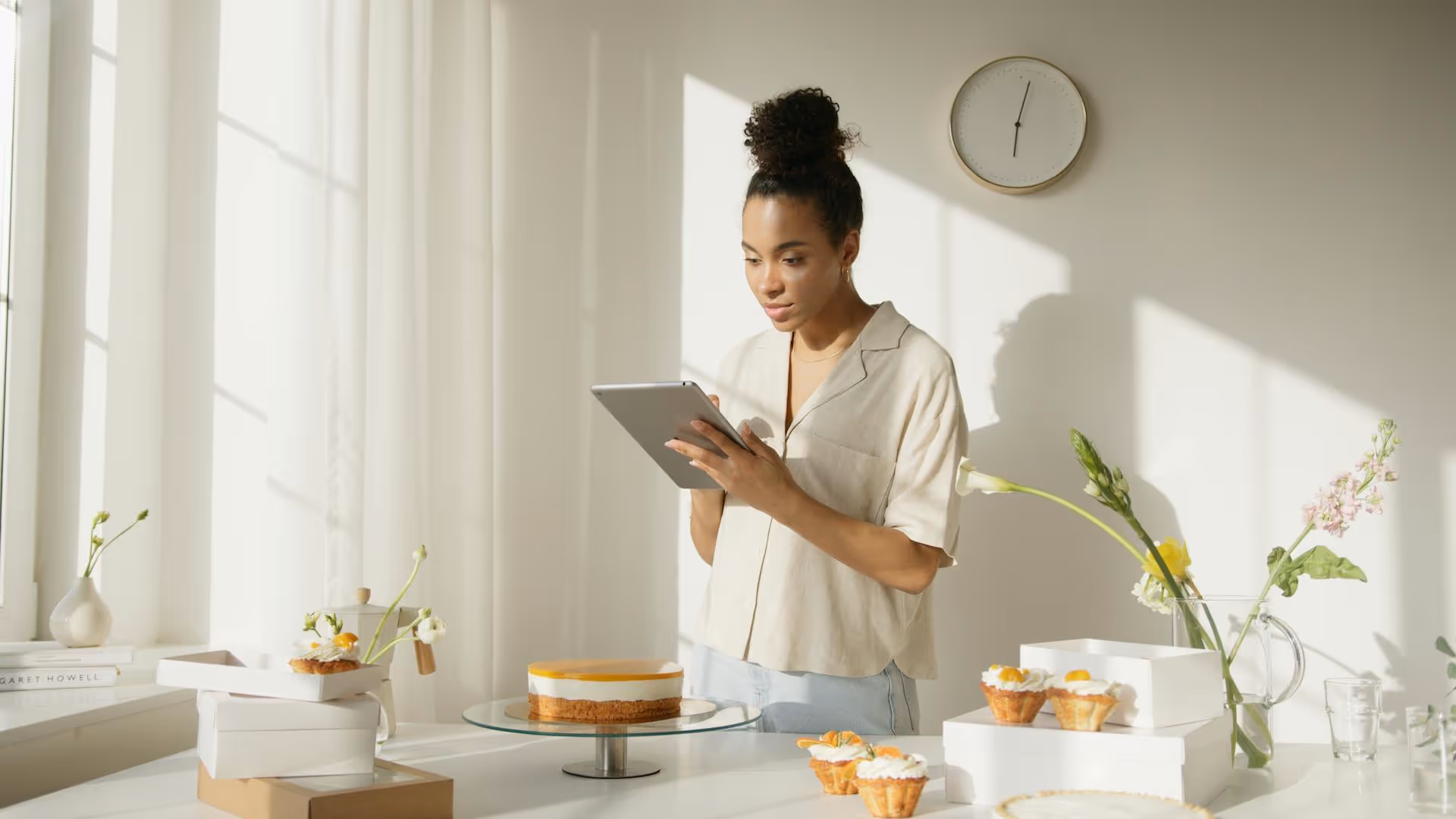 Woman in light shirt using a tablet near a table with a cake, cupcakes, and flowers in a bright kitchen.