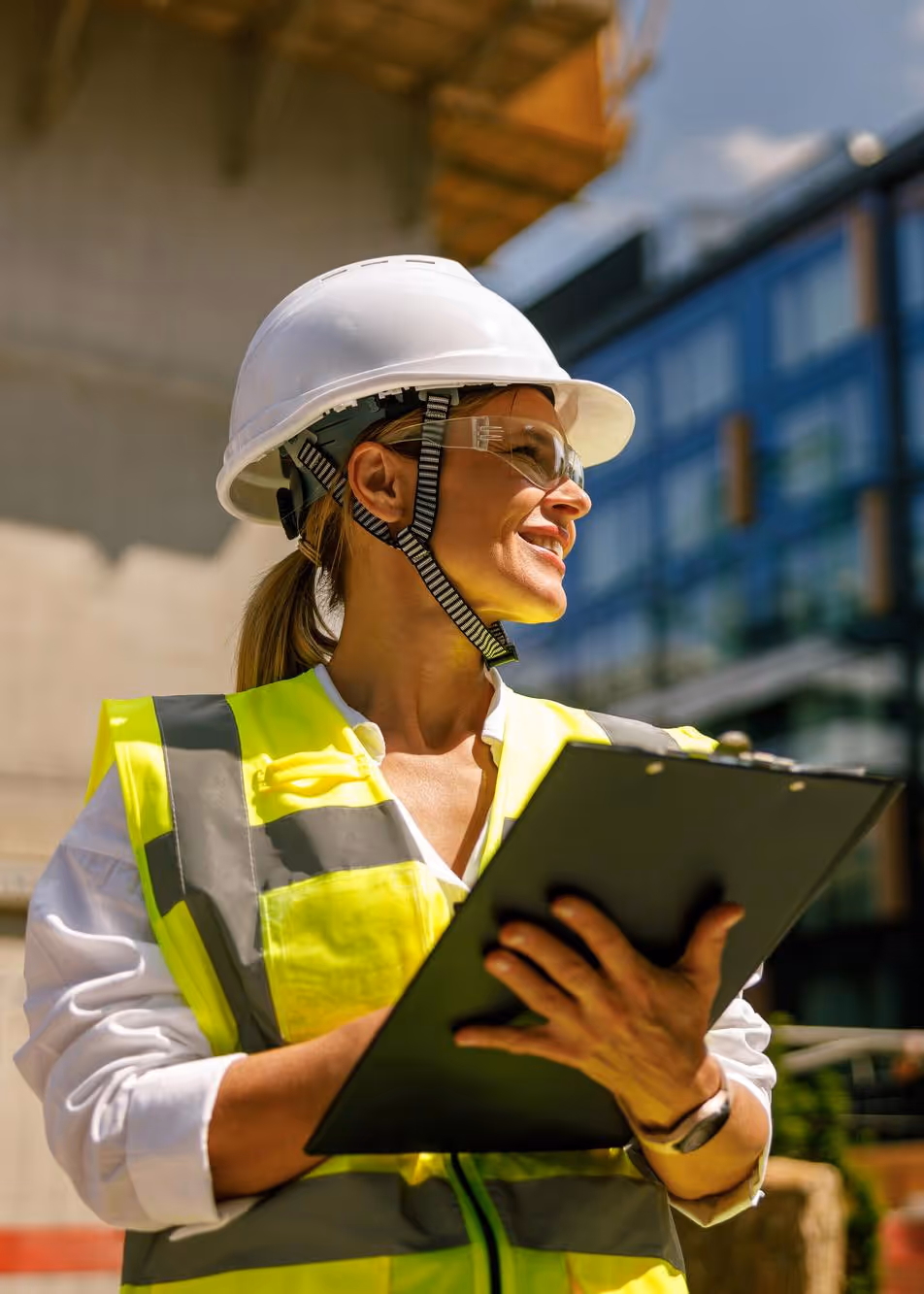 Woman construction worker in a hard hat and safety vest holding a clipboard outdoors.