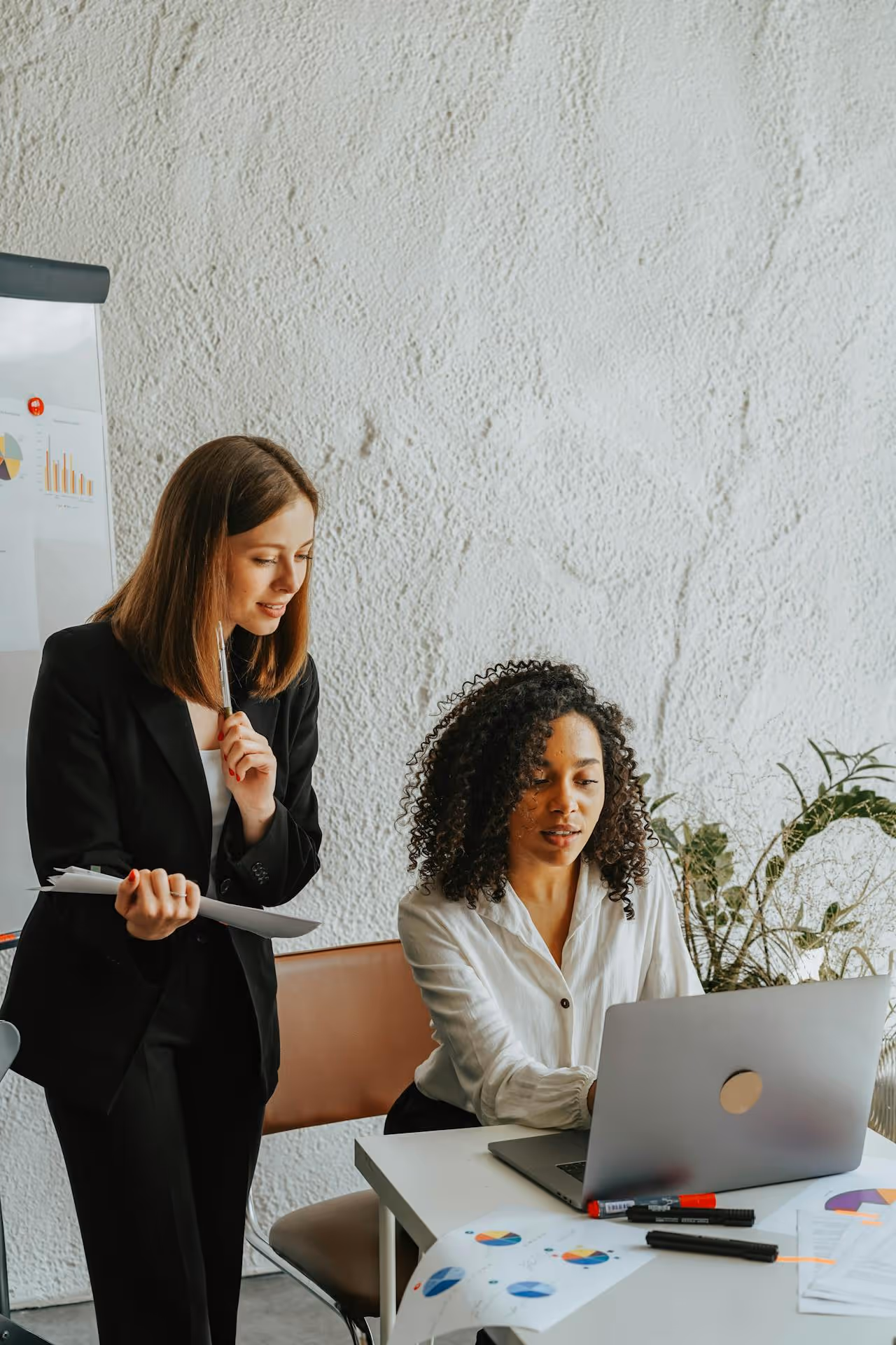 Two women collaborating in an office, one sitting at a laptop and the other standing holding papers and a pen.