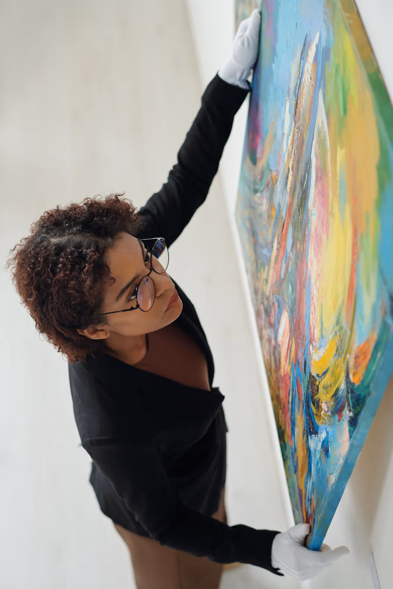 Woman wearing white gloves carefully hanging a colorful abstract painting on a wall.