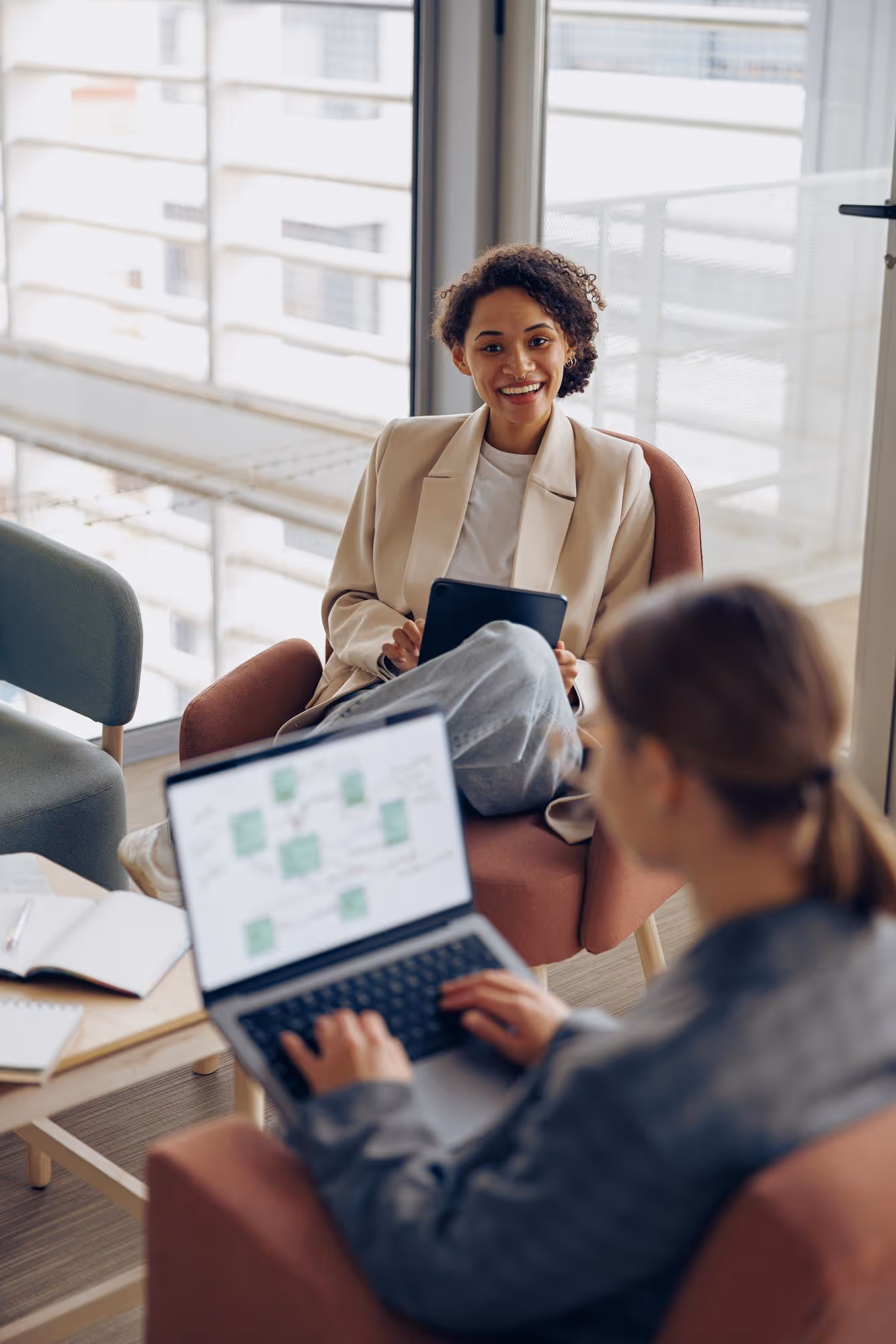 Two women in a modern office, one smiling and holding a tablet, the other working on a laptop with charts on screen.