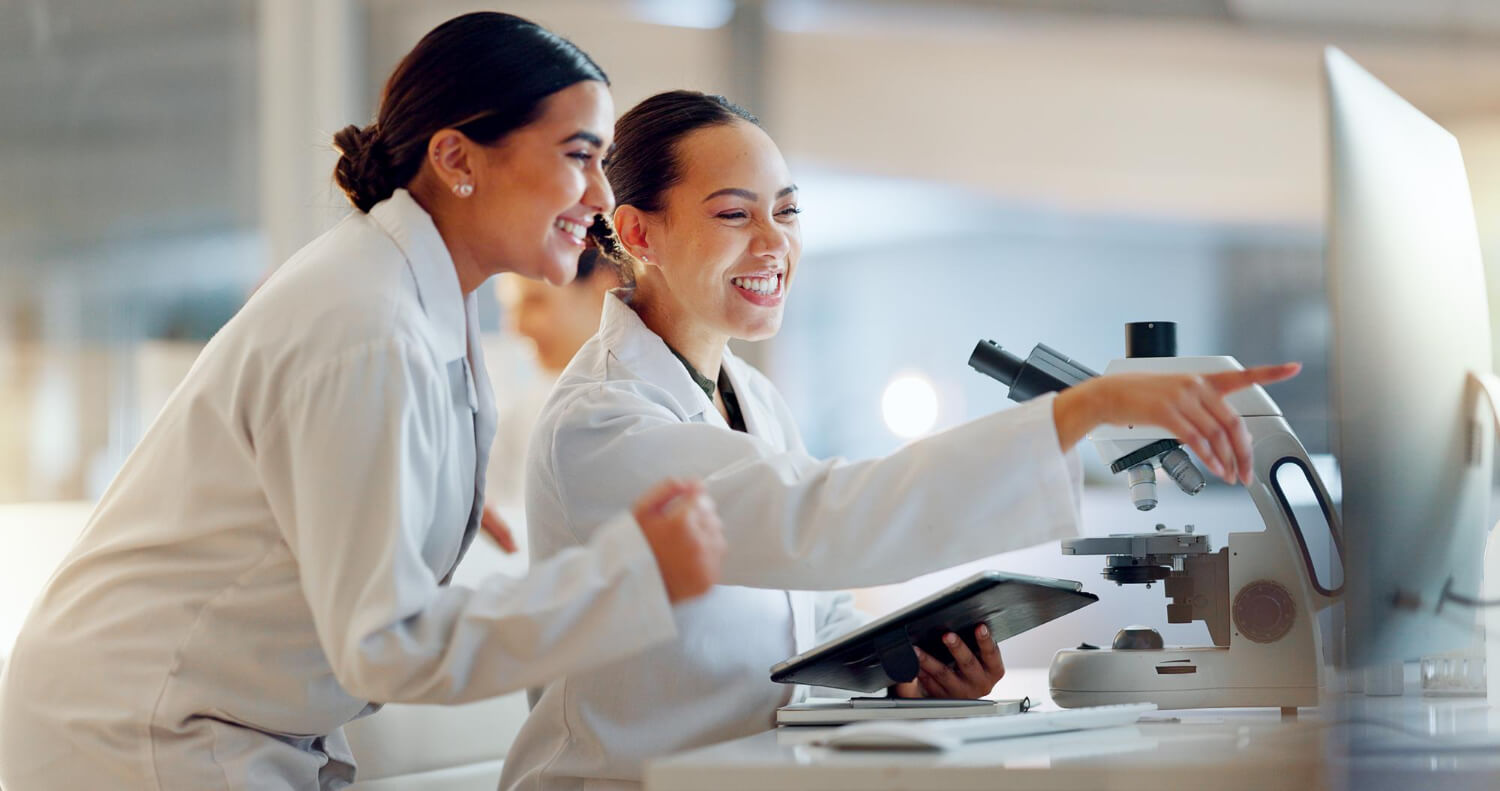 Two female scientists in lab coats smiling and pointing at a computer screen beside a microscope.
