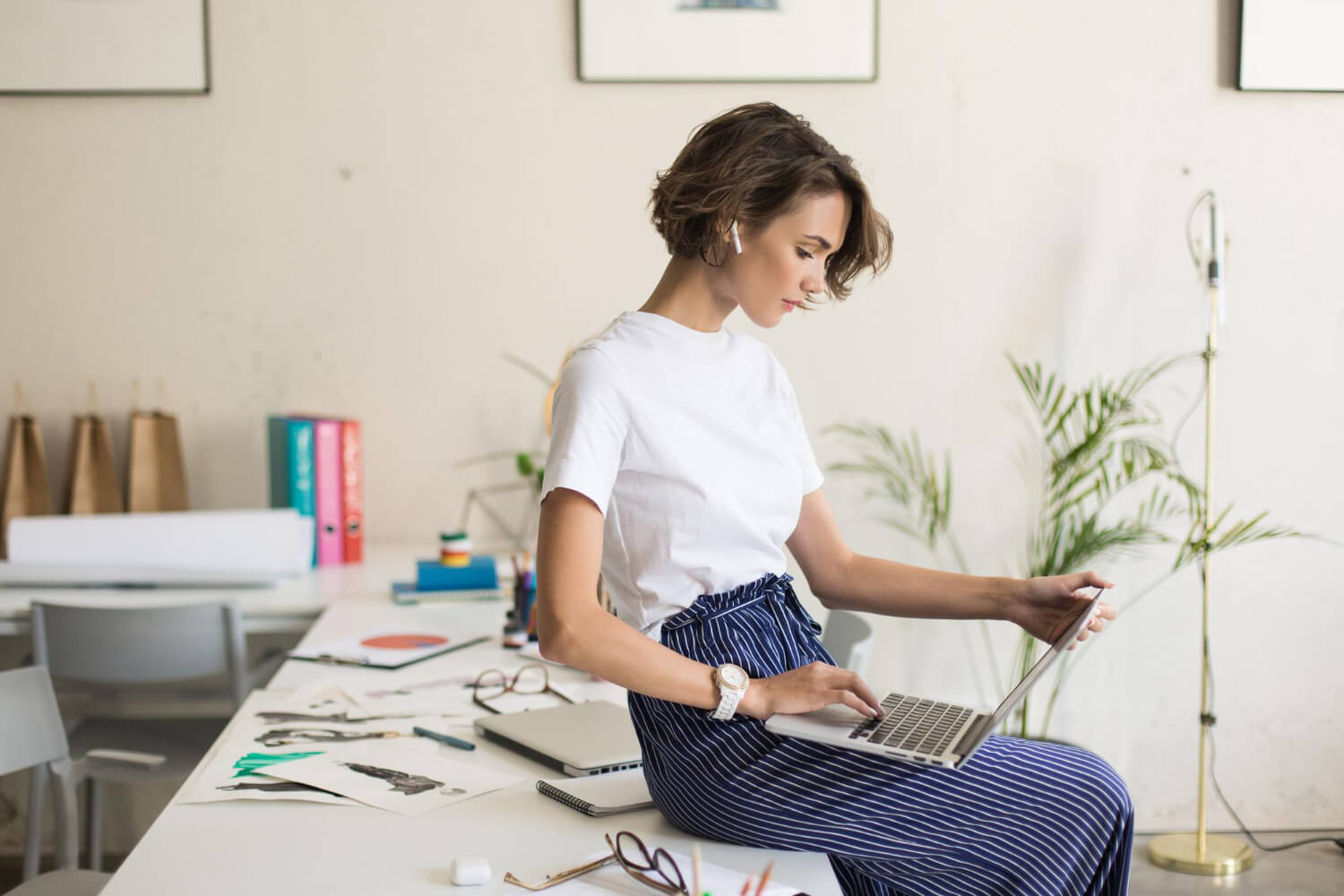 Woman sitting on a desk working on a laptop in a bright, modern office space.
