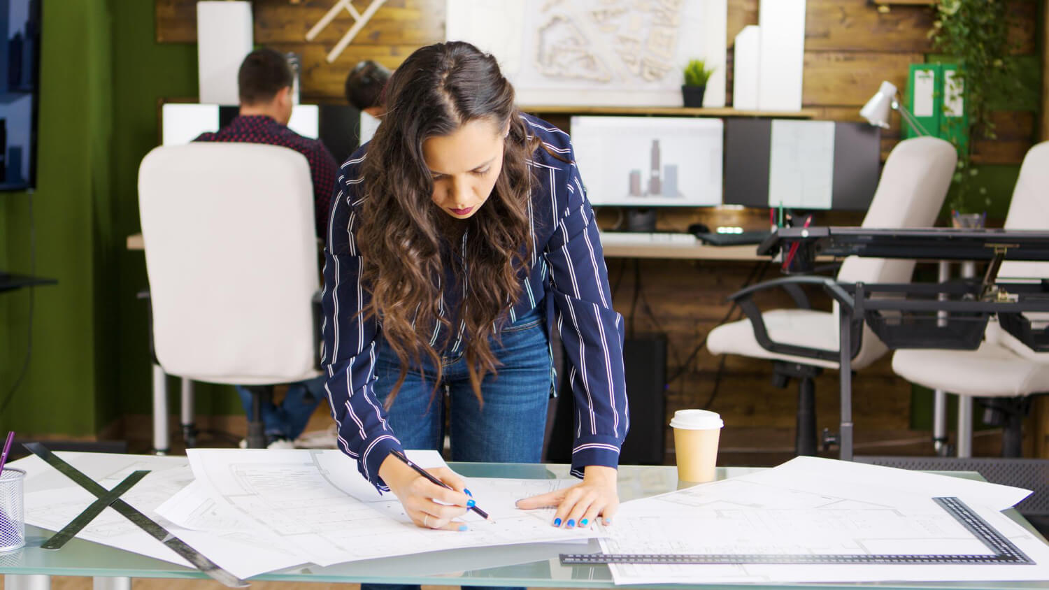 Woman with long curly hair leaning over a desk, drawing on architectural blueprints in an office.