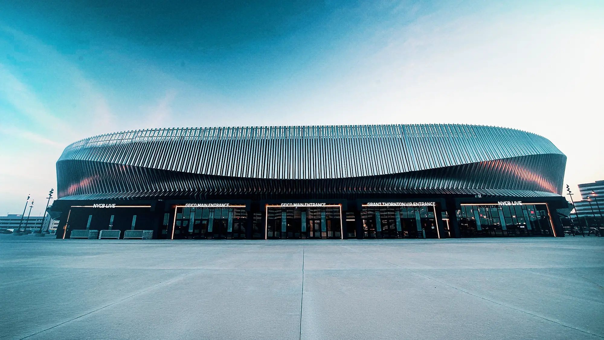 Wide low-angle view of a modern arena with vertical metal slats on its façade and multiple entrances labeled with illuminated signage, under a clear blue sky.