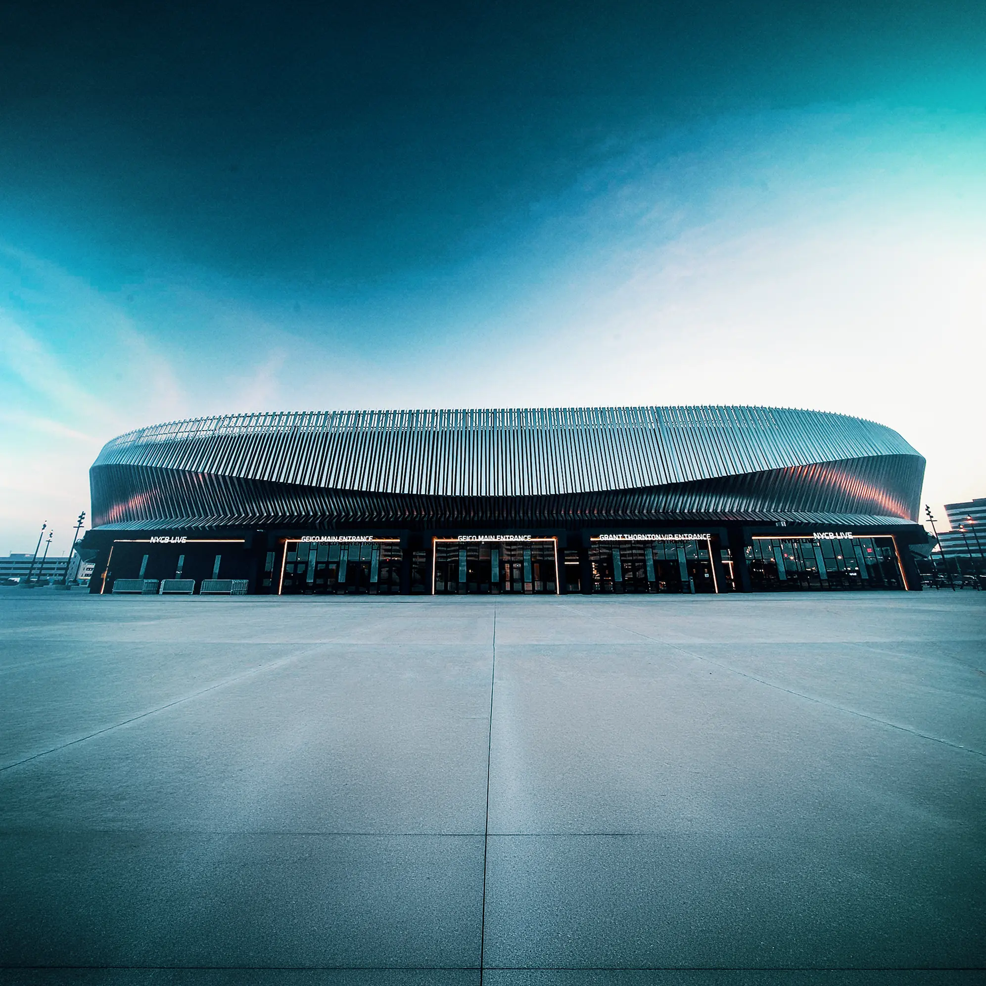 Modern arena or stadium with a distinctive corrugated metal facade, wide entrance doors labeled with signs including 'GEICO MAIN ENTRANCE', 'GRANT THORNTONVIP ENTRANCE', and 'NYCB LIVE', under a twilight sky.