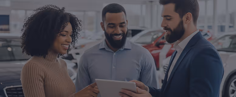 Man and woman smiling while looking at a tablet held by a third man in a suit inside a car showroom.