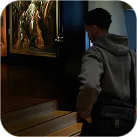 Person in a gray hoodie and black crossbody bag viewing a framed painting on a dark wall in a dimly lit room.