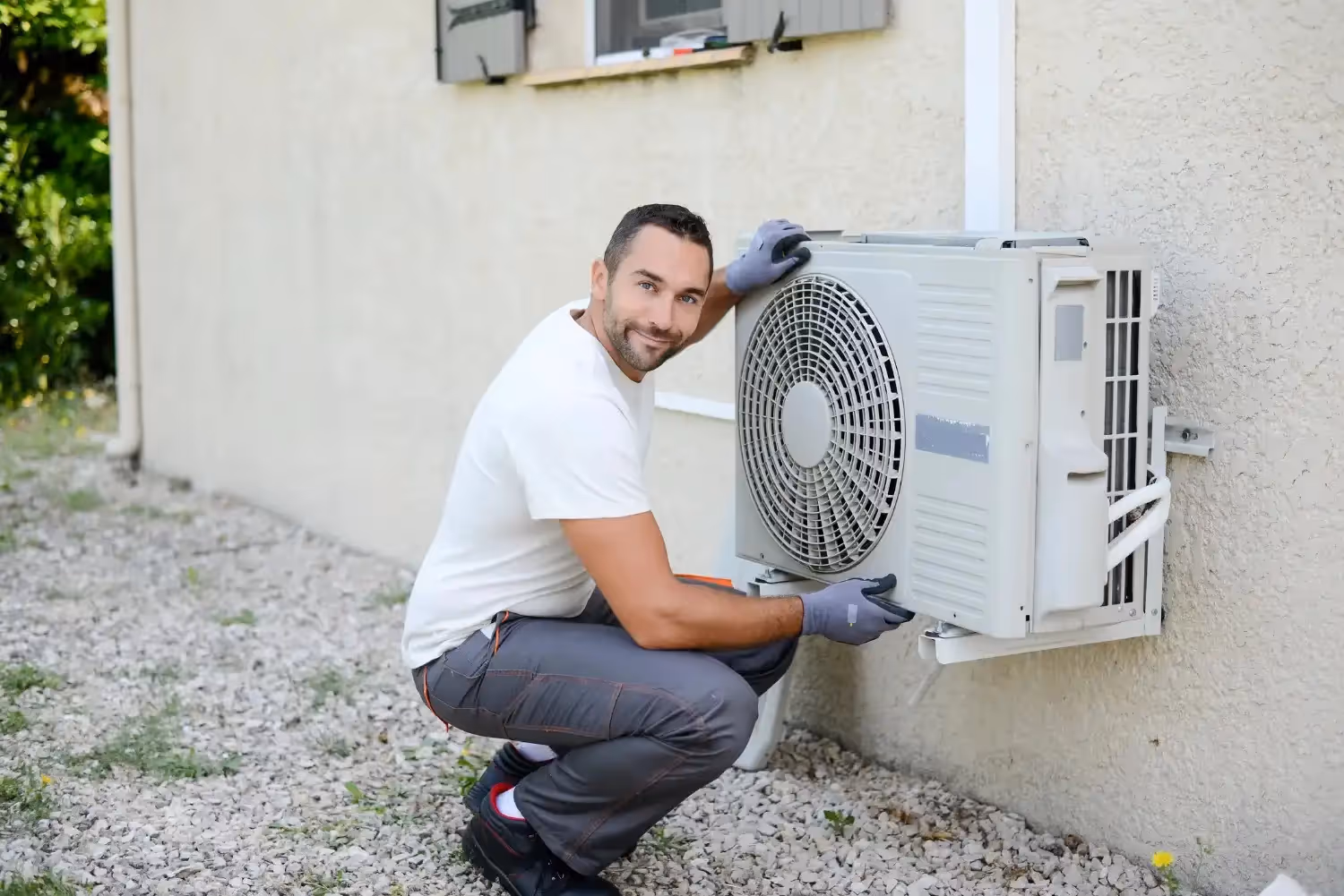 A smiling HVAC technician wearing gray gloves and a white shirt poses next to an outdoor AC unit