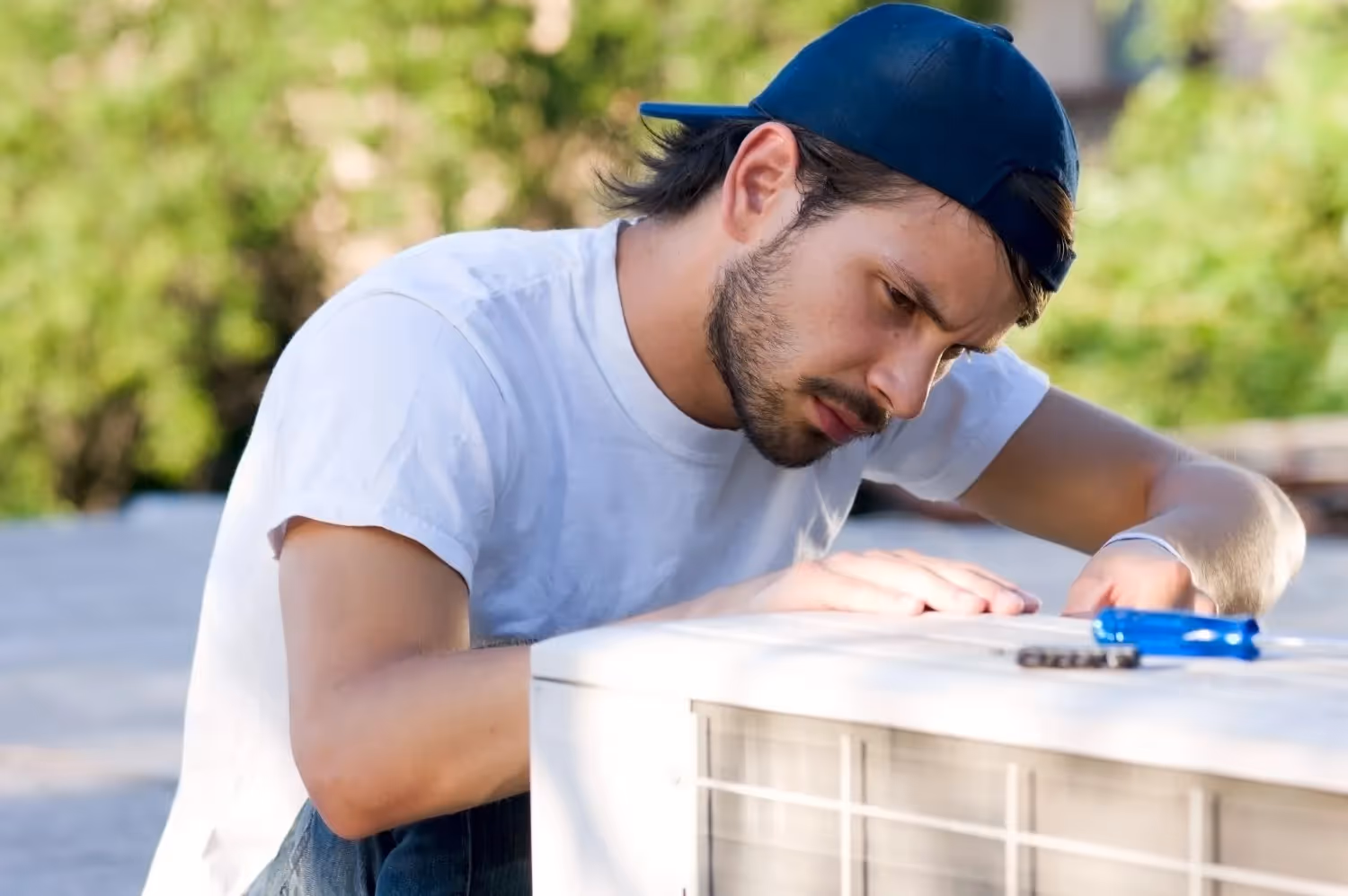 A technician wearing a tool belt and a cap is kneeling and repairing an outdoor heat pump unit next to a modern wooden wall