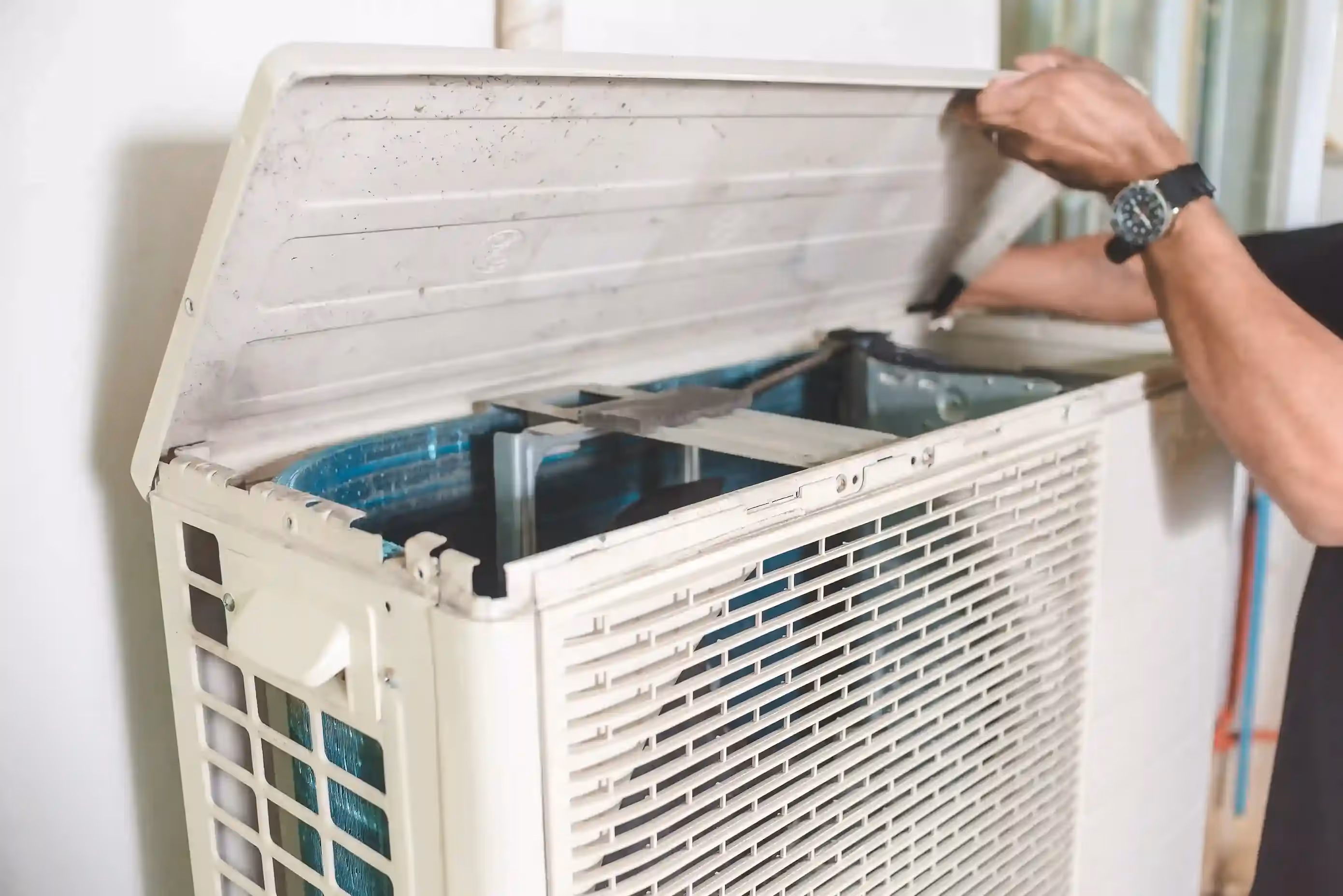 A close-up of a person's hands lifting the lid off an outdoor heat pump unit, exposing the internal components.