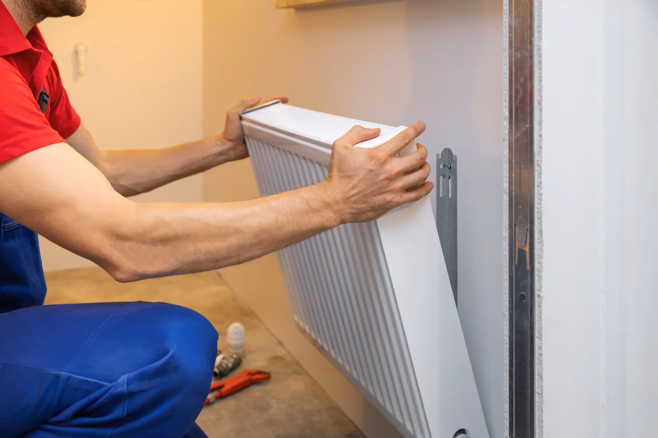 A person in blue overalls holds a white heating radiator, preparing to mount it on a wall bracket.