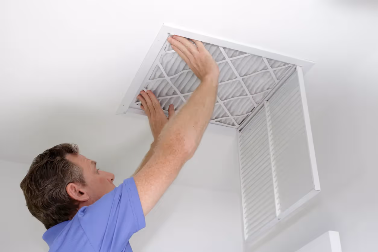 A man reaches up to a ceiling air vent, pulling down the grille to access the filter