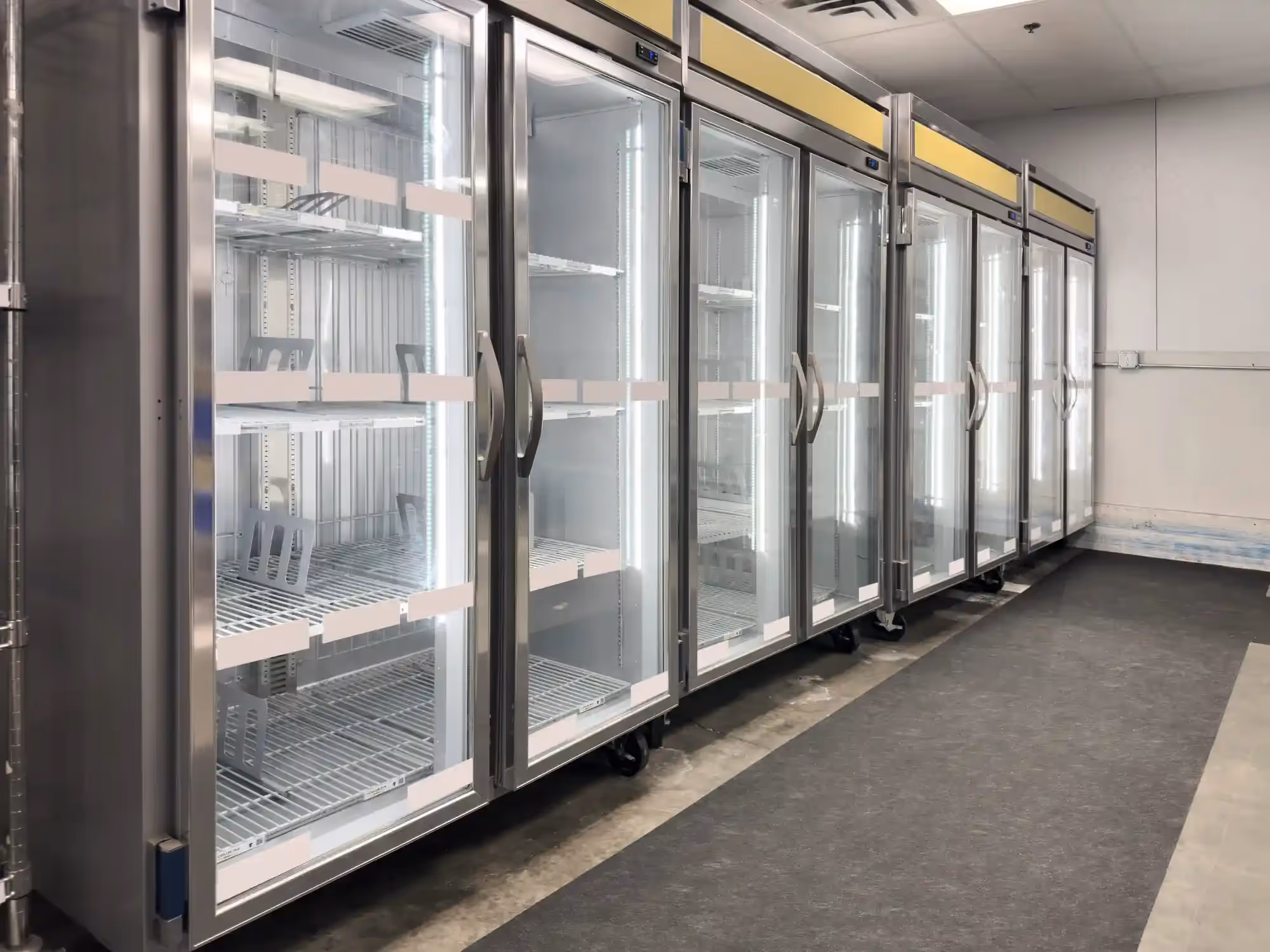 A row of new, empty commercial reach-in refrigerators with glass doors, illuminated interiors, and wire shelves, ready for stocking.