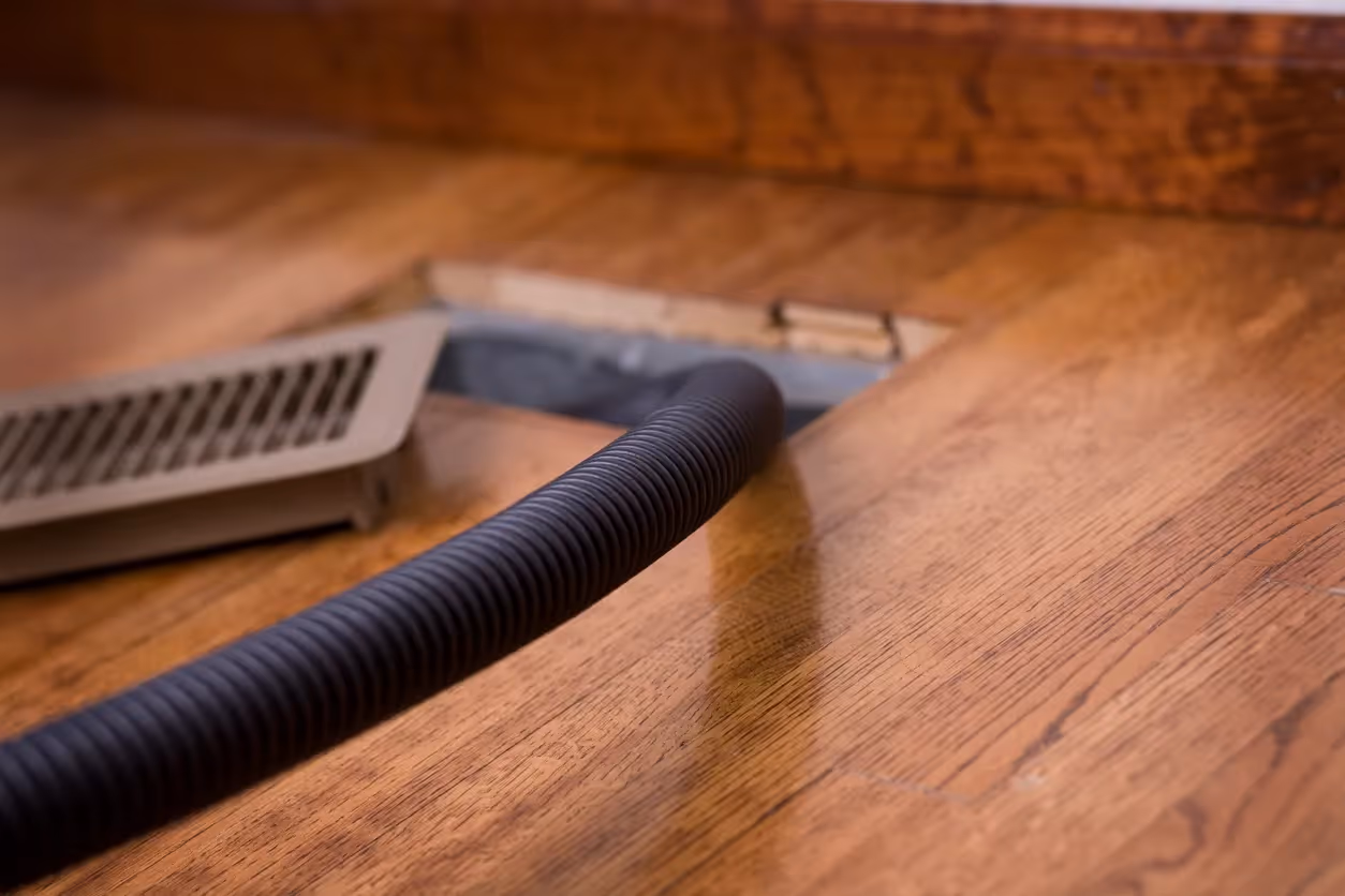A close-up of a vacuum hose cleaning a dusty air vent on a hardwood floor