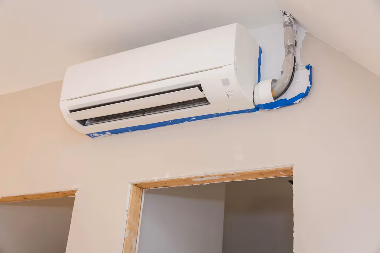 A white mini-split unit is mounted on a beige wall with a pipe wrapped in blue tape entering the wall