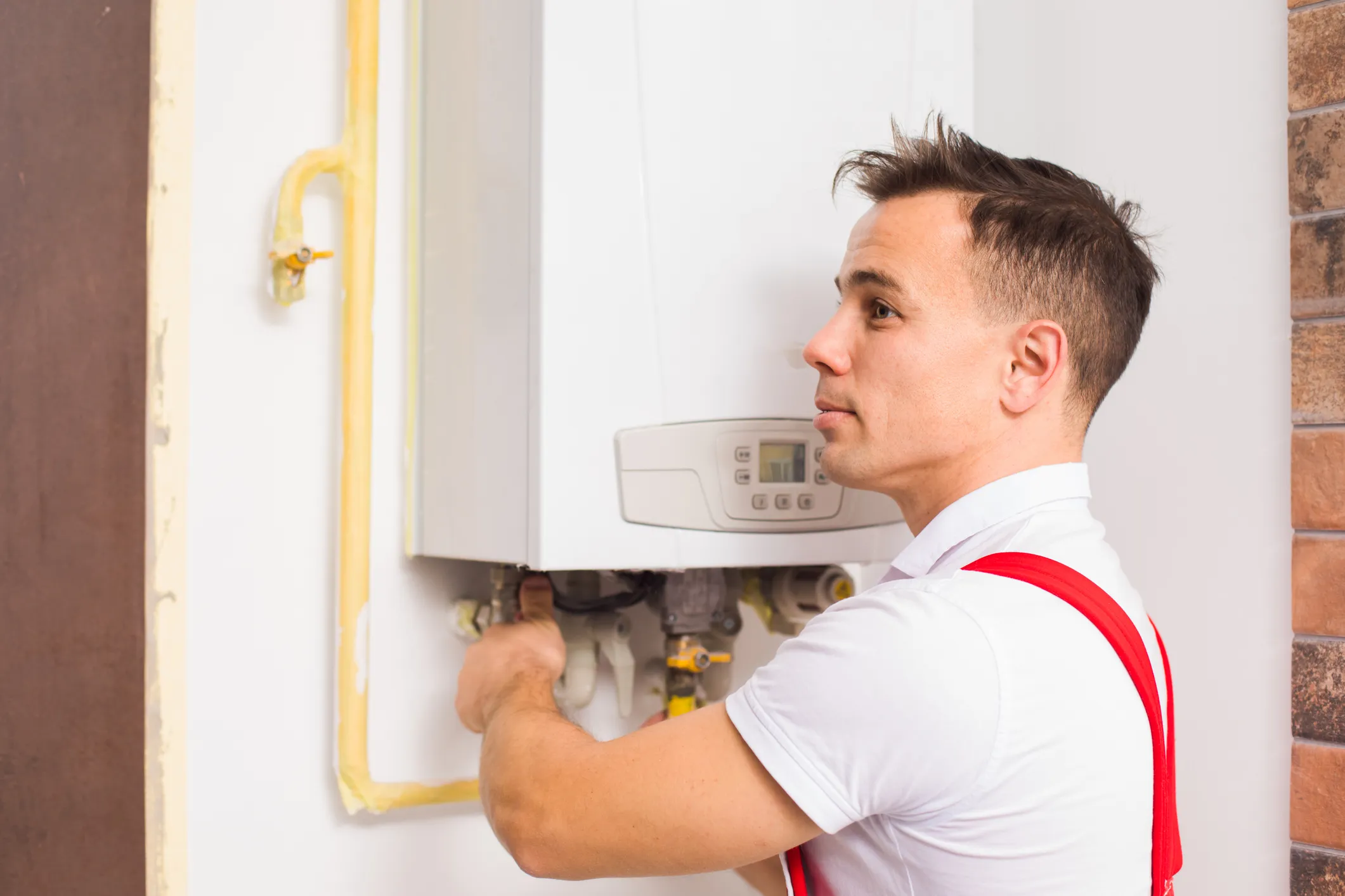 Plumber in red overalls adjusting pipes at the bottom of a white boiler.