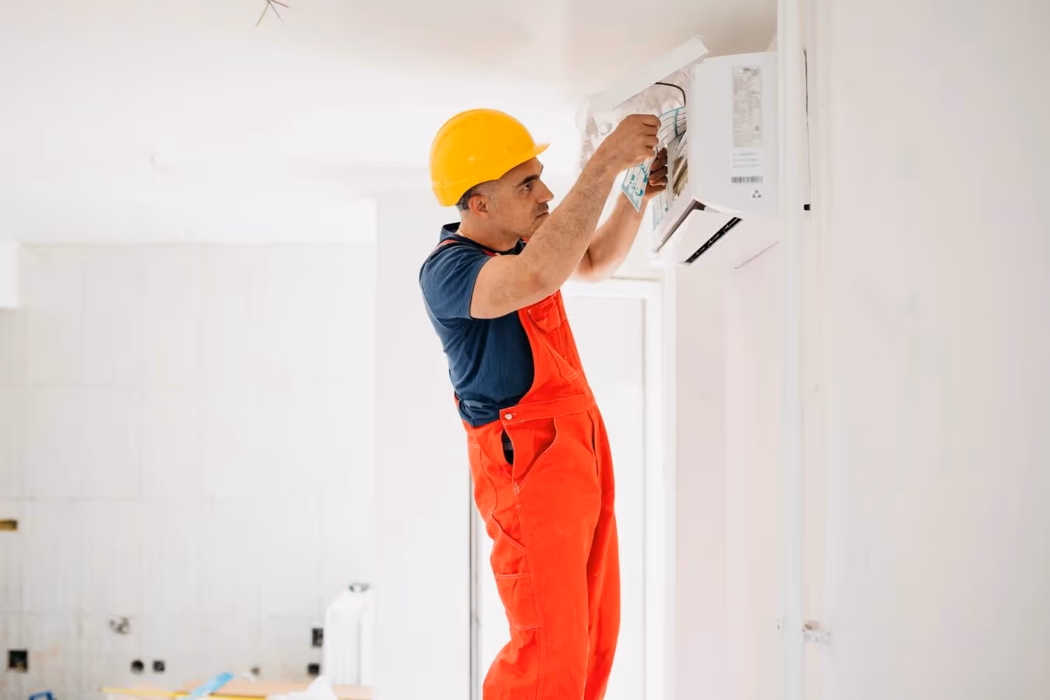A professional technician, wearing a yellow hard hat and orange overalls over a blue shirt, is installing or servicing a mini-split air conditioner. He's standing and looking up, with his hands inside the unit's casing, connecting wires. The unit is mounted on a white wall in a bright, sparsely furnished room, possibly a new construction or a renovation.