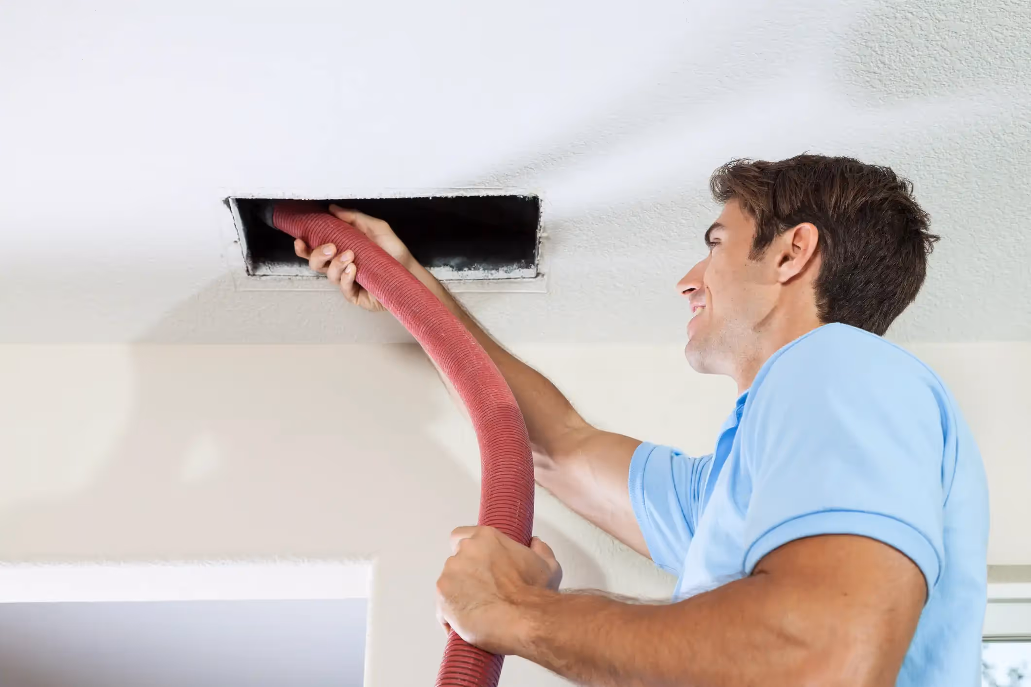 A smiling technician holds a red flexible vacuum hose, inserting it into a rectangular ceiling air vent to clean the duct.
