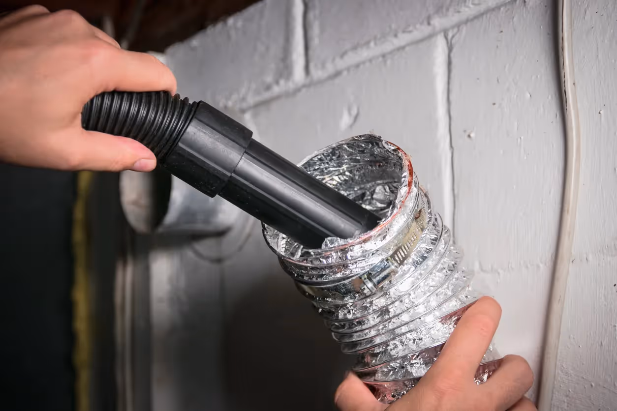 A hand holds a vacuum hose nozzle, inserting it into a flexible metallic air duct to clean the interior.