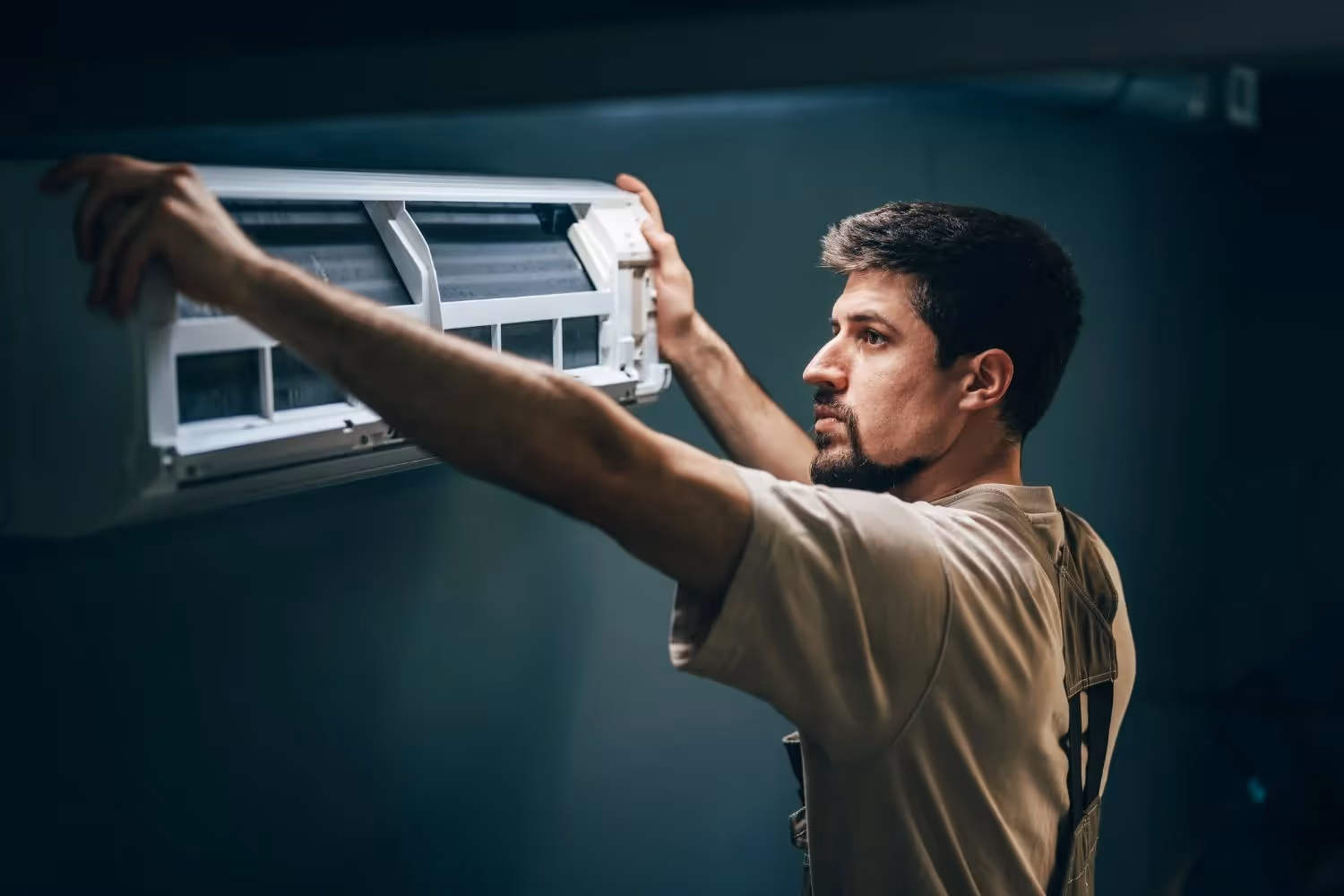 A man with a beard, wearing a tan t-shirt and work vest, is installing the indoor unit of a mini-split air conditioner on a dark gray wall. He is holding the unit with both hands, looking at it intently from the side as he positions it.