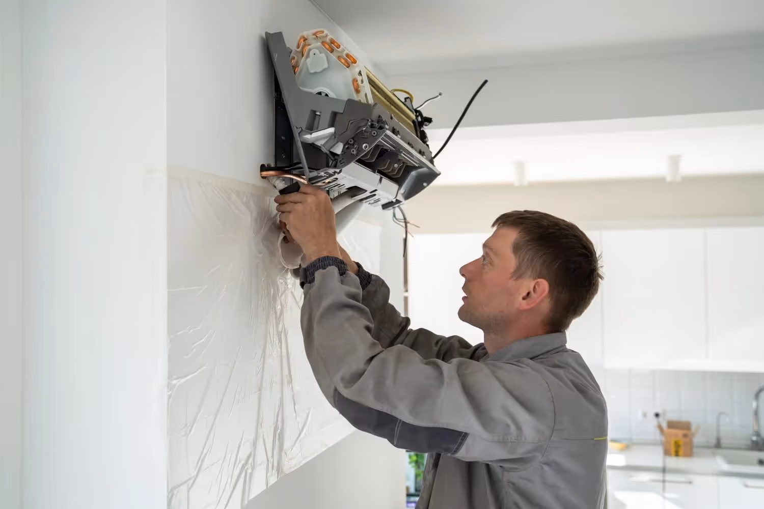 A professional HVAC technician is connecting the refrigerant lines to the indoor unit of a mini-split air conditioner. The technician, a man with short brown hair, is wearing a gray long-sleeved uniform. He's focused on the pipes, with one hand holding a tool and the other guiding the connection. The indoor unit is mounted on a white wall, with the plastic cover removed to expose the internal components. A protective plastic sheet is taped to the wall below the unit to prevent damage or mess. In the background, there's a modern kitchen with white cabinets.