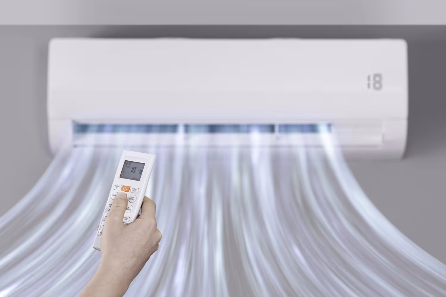 A hand holds a remote control, operating a wall-mounted air conditioner that is blowing air.