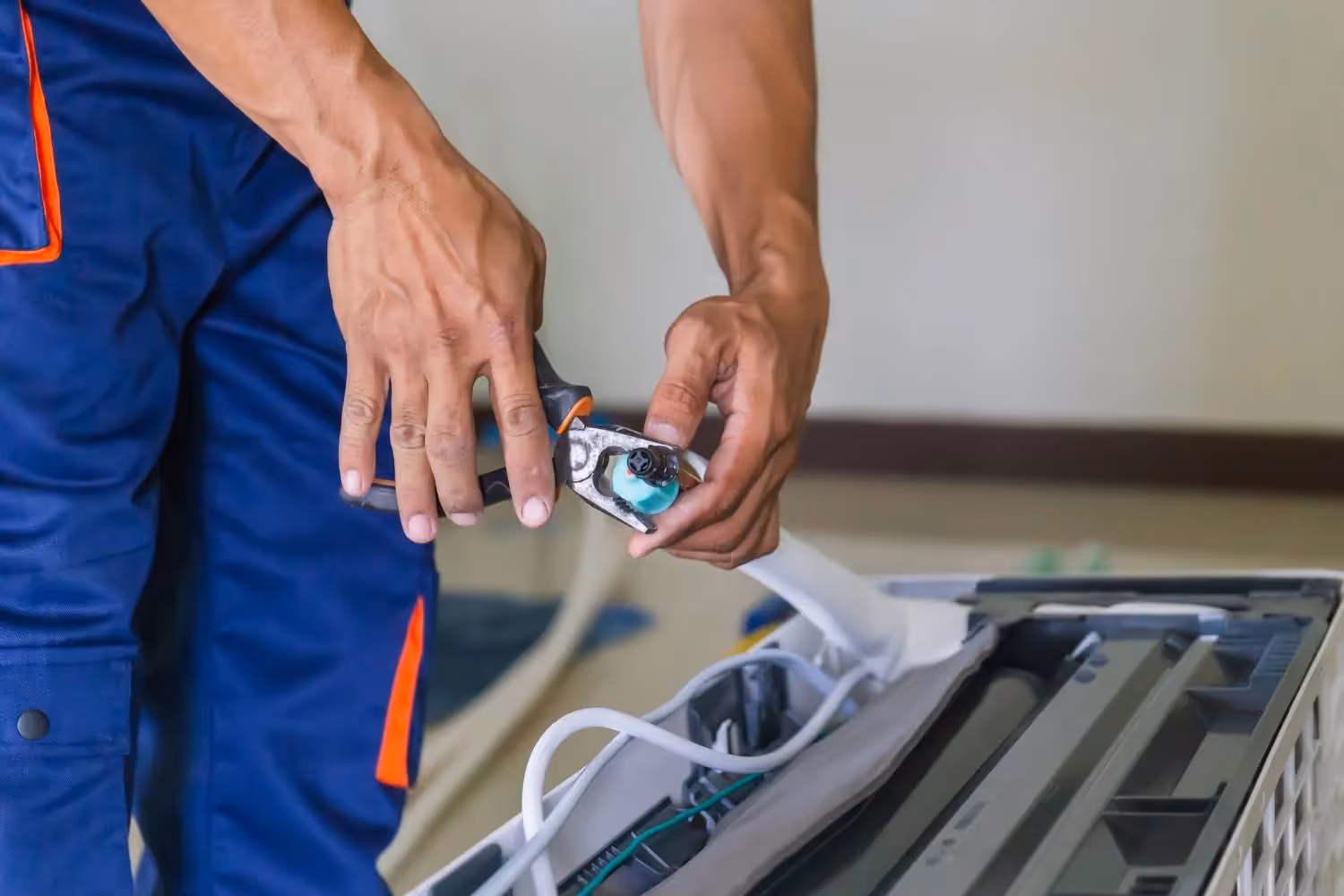 A close-up shot of a technician's hands, in blue and orange uniform pants, using a pair of pliers to work on a small part of a mini-split air conditioner. The technician is holding the pliers in his right hand and the part in his left, with the AC unit's internal components visible in the foreground. The background is out of focus, suggesting an indoor setting.