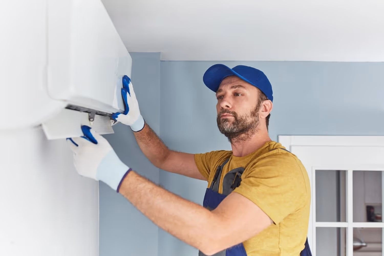 A male HVAC technician with a beard is working on a mini-split air conditioner unit mounted high on a wall. He is wearing a yellow shirt, a blue baseball cap, blue overalls, and white and blue gloves. He's focused on the unit, holding a white panel with both hands, either installing or removing it. The room has light blue walls and a white ceiling.
