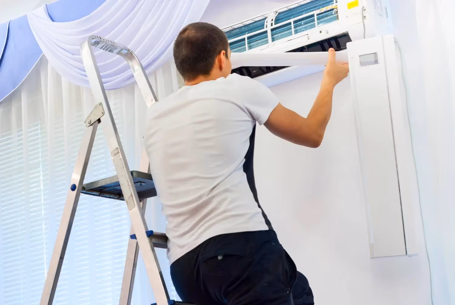 A person in a white t-shirt and dark pants is standing on a stepladder, facing away from the camera, as they clean or work on the internal fan of a mini-split air conditioner unit. The person's left hand is holding the fan while their right hand is inserting or removing a part from the unit. The AC unit is mounted on a white wall above a window with blinds and blue curtains.