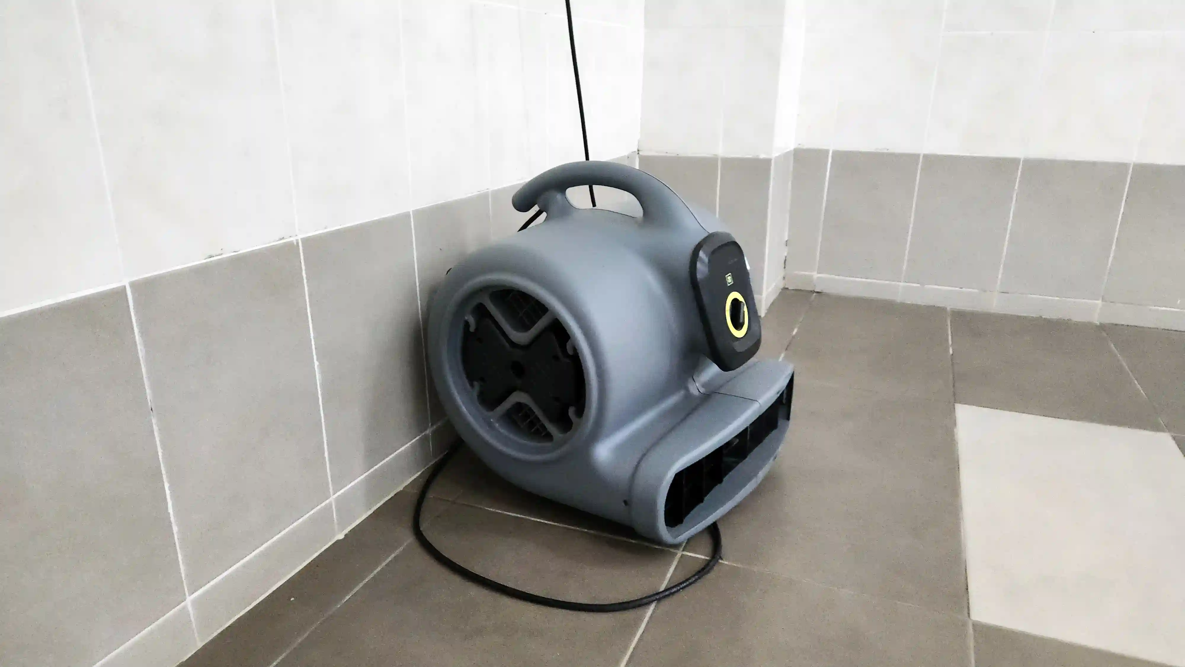 A close-up of a gray, portable industrial fan with a handle on top. The fan has a yellow circular button and is sitting on a tiled floor in the corner of a room with tiled walls.