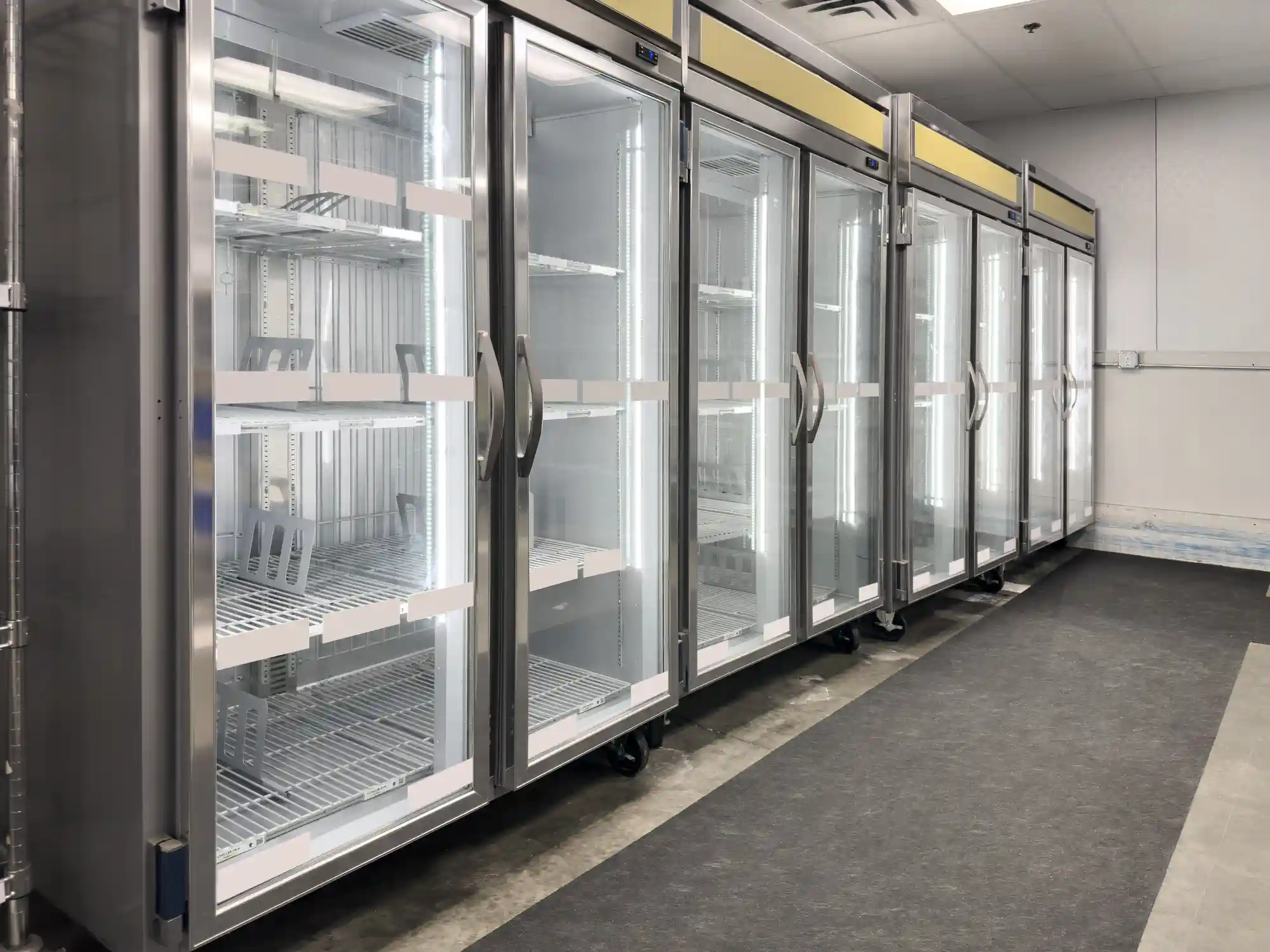 A line of large, empty, stainless steel commercial refrigerators with glass doors is visible in a room. The refrigerators have multiple wire racks inside and are lit with bright interior lighting. They are on a gray floor, with a darker gray mat running in front of them.
