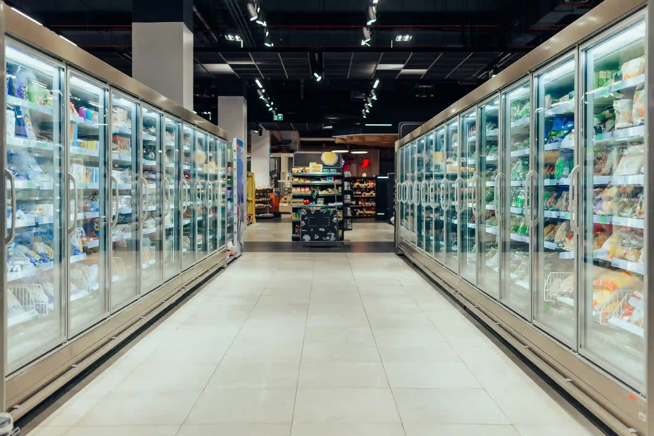 A wide-angle, symmetrical shot shows an aisle in a grocery store, lined on both sides with large commercial refrigerators with glass doors. The refrigerators are filled with frozen food products. The floor is made of light-colored tiles and leads to a blurred view of other store aisles in the distance. The ceiling has exposed dark pipes and lights.
