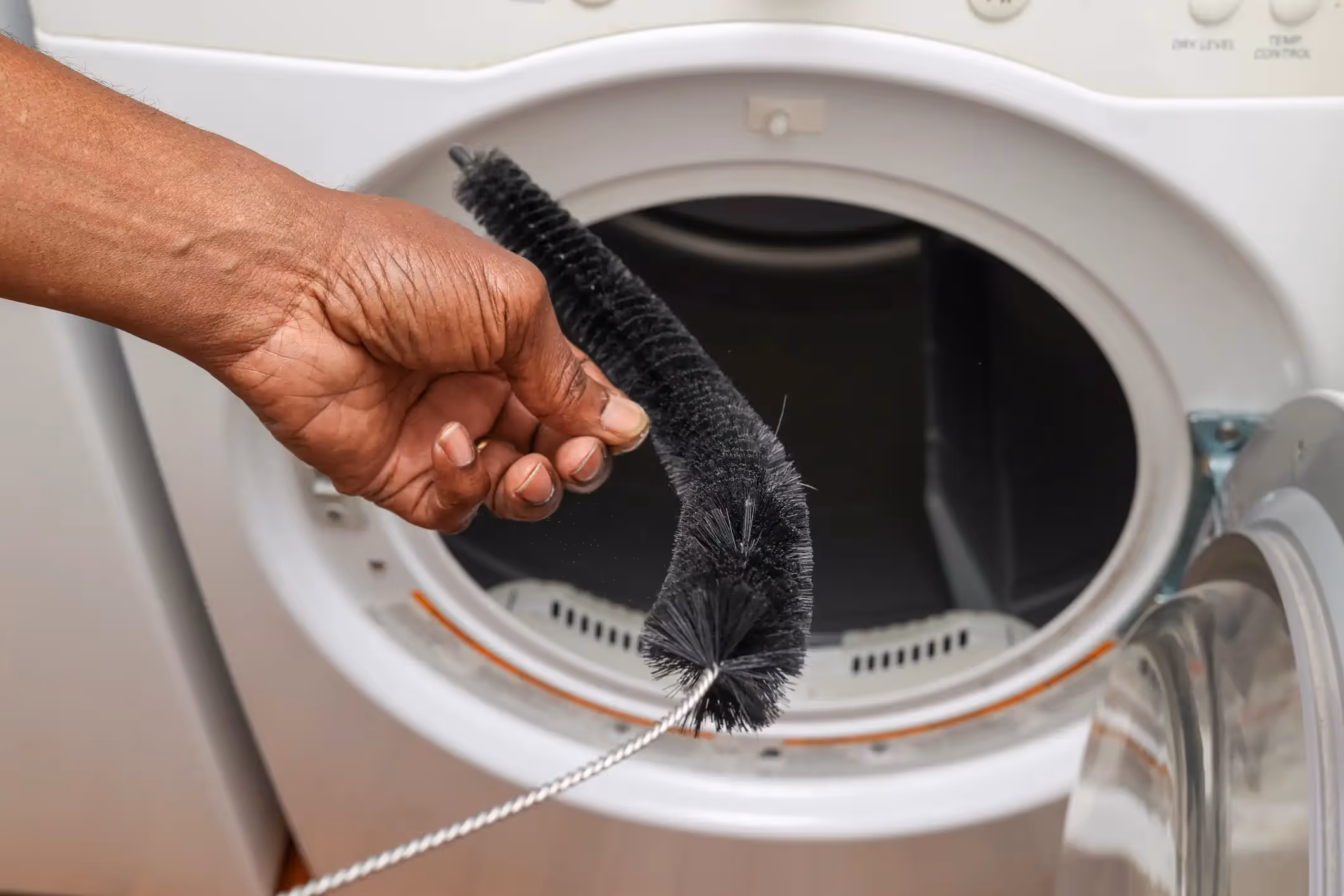 A person with brown skin holds a long, flexible black brush with a metal wire handle, used for cleaning lint. The brush is positioned in front of the open door of a white clothes dryer. The interior of the dryer's drum and the lint screen are visible.