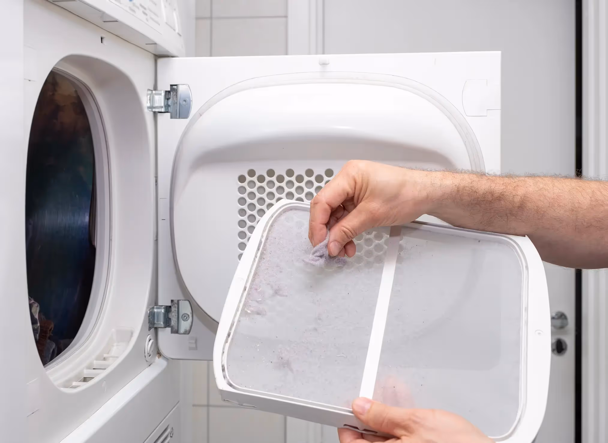 A person's hands are shown removing a handful of gray lint from the lint filter of a white clothes dryer. The filter is a white plastic frame with a mesh screen, and the open door of the dryer is visible in the background.