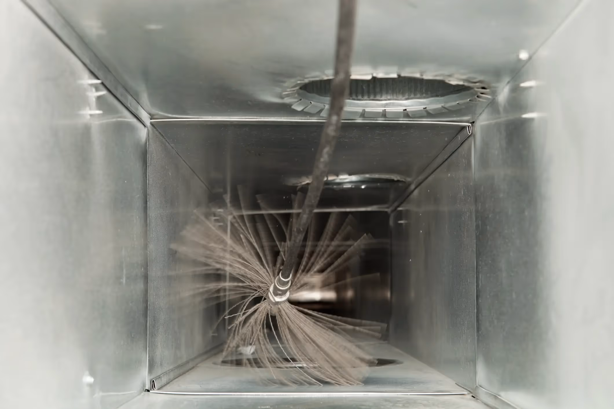 A high-angle view looking down into a square metal air duct. A long, thin rod with a spinning, gray wire brush at its end is visible inside the duct, centered in the shot. The brush appears to be in motion, and a circular opening is seen in the ceiling of the duct above it.