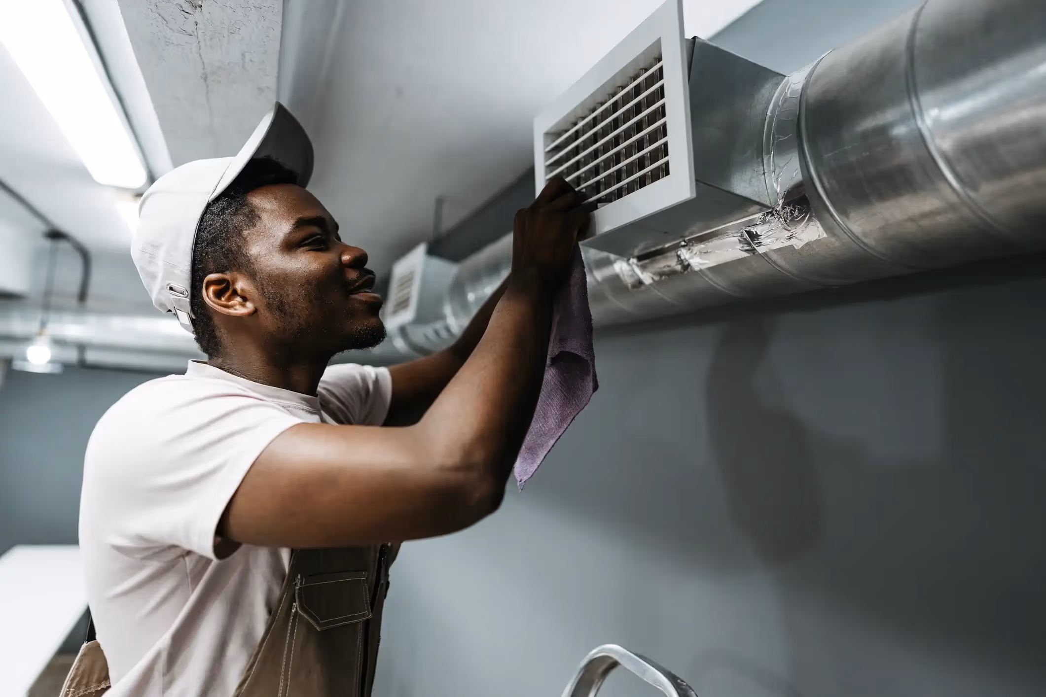 A male technician with a light-colored baseball cap and a light pink shirt is smiling while using a purple cloth to clean a rectangular air vent grill attached to a large, shiny, silver duct pipe. The pipe and vent are mounted on a gray wall in what appears to be a commercial or industrial building.