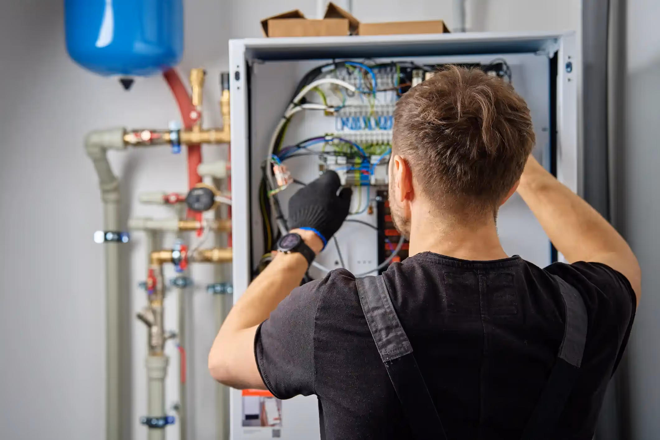 A technician wearing black gloves and a black t-shirt with grey overalls is working on the internal components of a white electrical box. The box has a panel open, revealing a complex network of wires, circuit boards, and connectors.