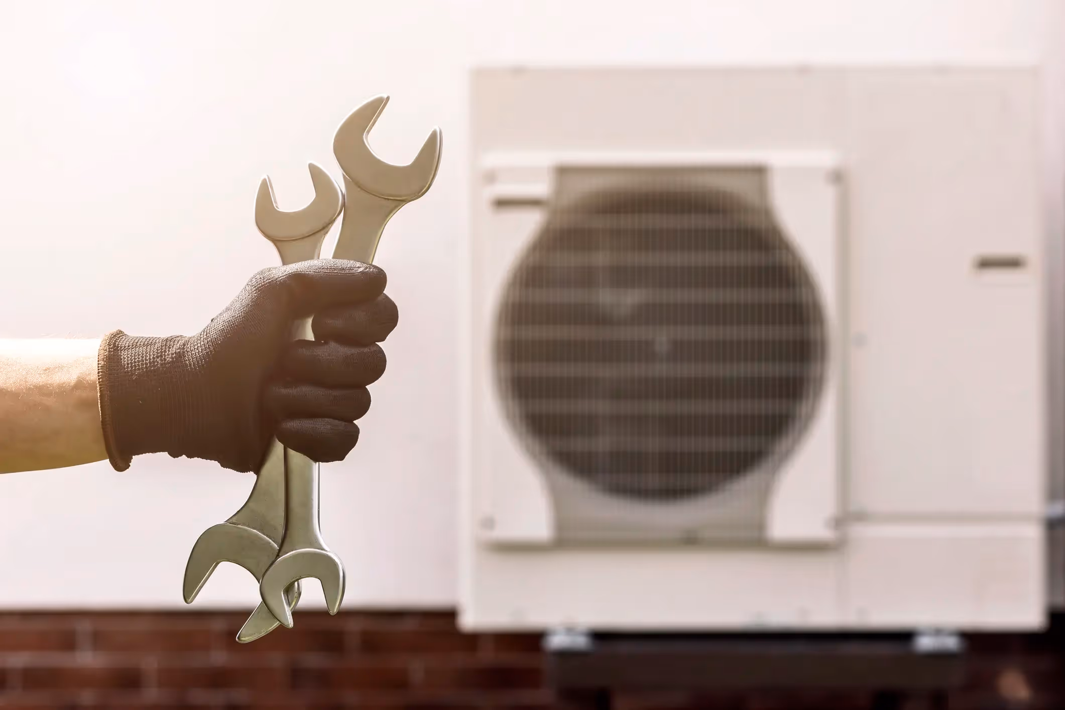 A close-up shot of a hand in a black glove holding a pair of large wrenches. The hand is positioned in the foreground, slightly out of focus. In the background, a white outdoor heat pump unit with a circular fan grille is visible, slightly blurred.