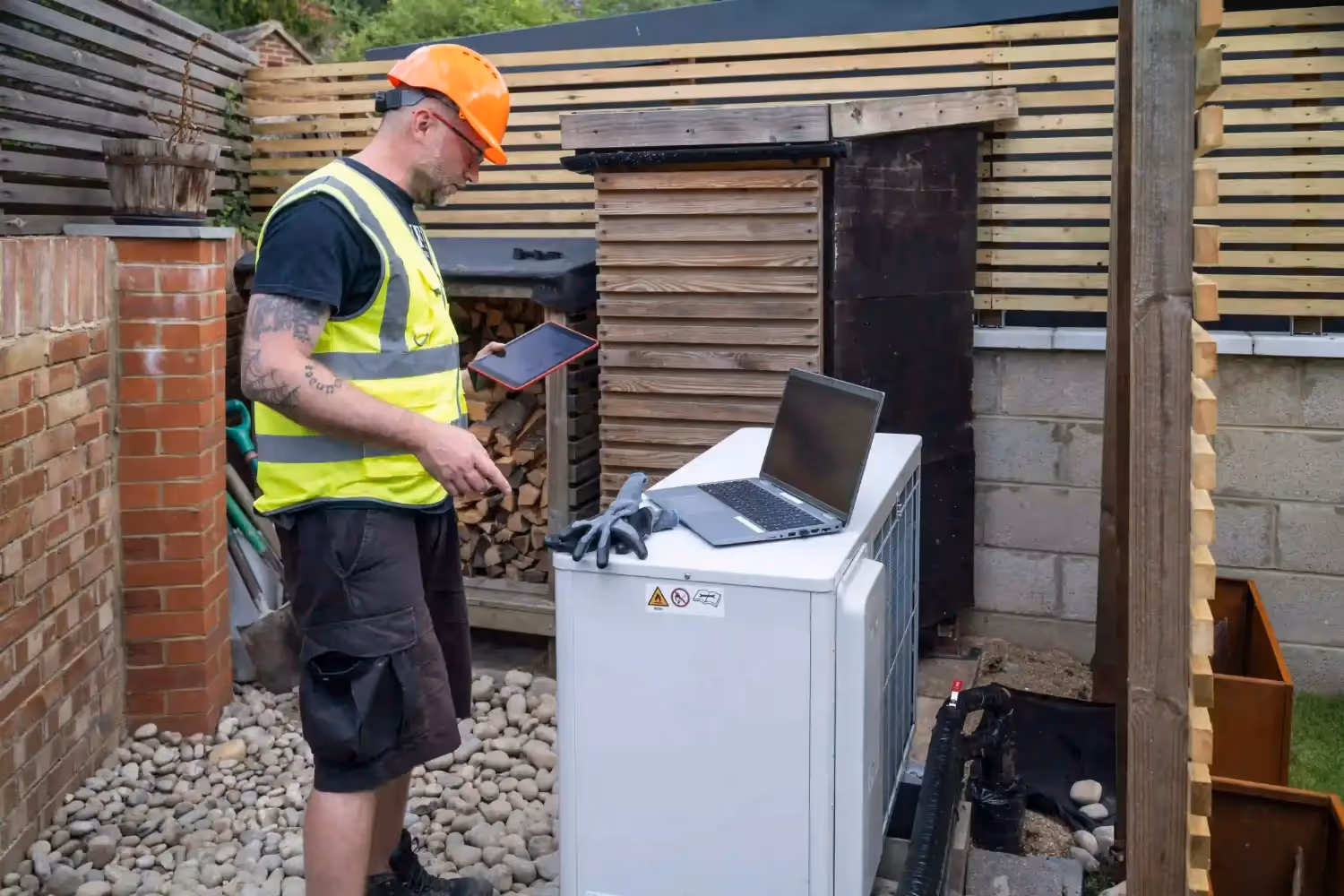 A male technician wearing an orange hard hat and a high-visibility yellow vest is standing in a backyard, working on a white outdoor heat pump unit. He is holding a tablet in one hand and looking down at a laptop that is resting on top of the unit. The background shows a brick wall, wood-slatted fencing, and a pile of small gray rocks on the ground.