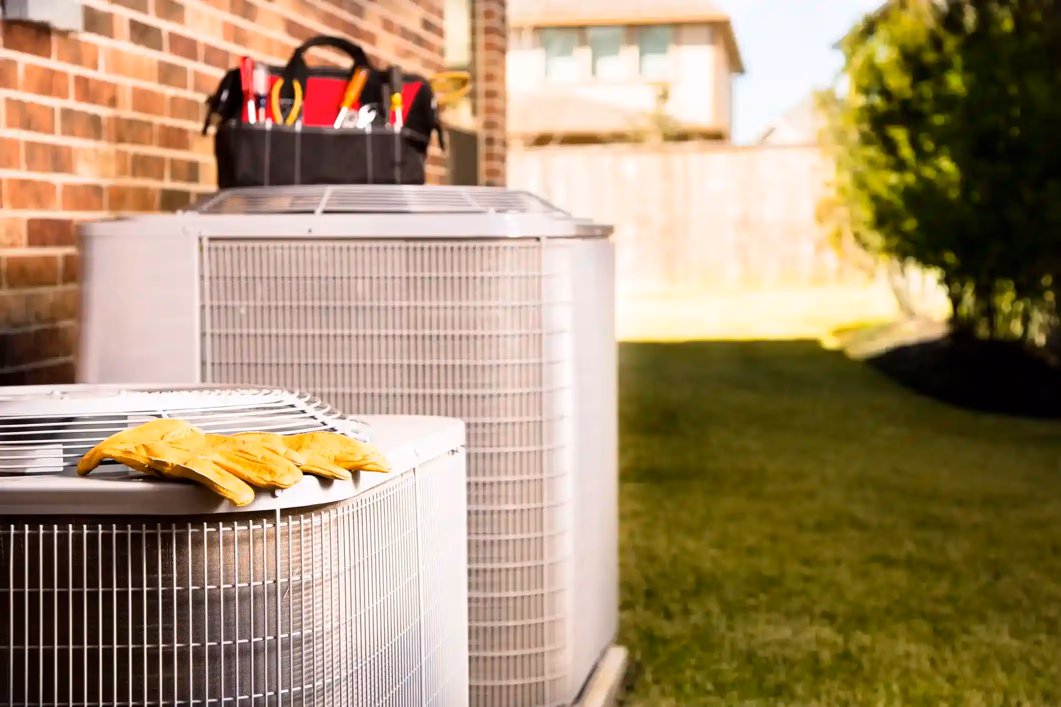 pair of yellow work gloves rests on top of a white outdoor air conditioning or heat pump unit, which is positioned in front of a red brick wall. In the background, a second, larger unit has a black toolbox filled with tools on top of it. The scene is outdoors on a grassy lawn with a blurred background of a house and fence.