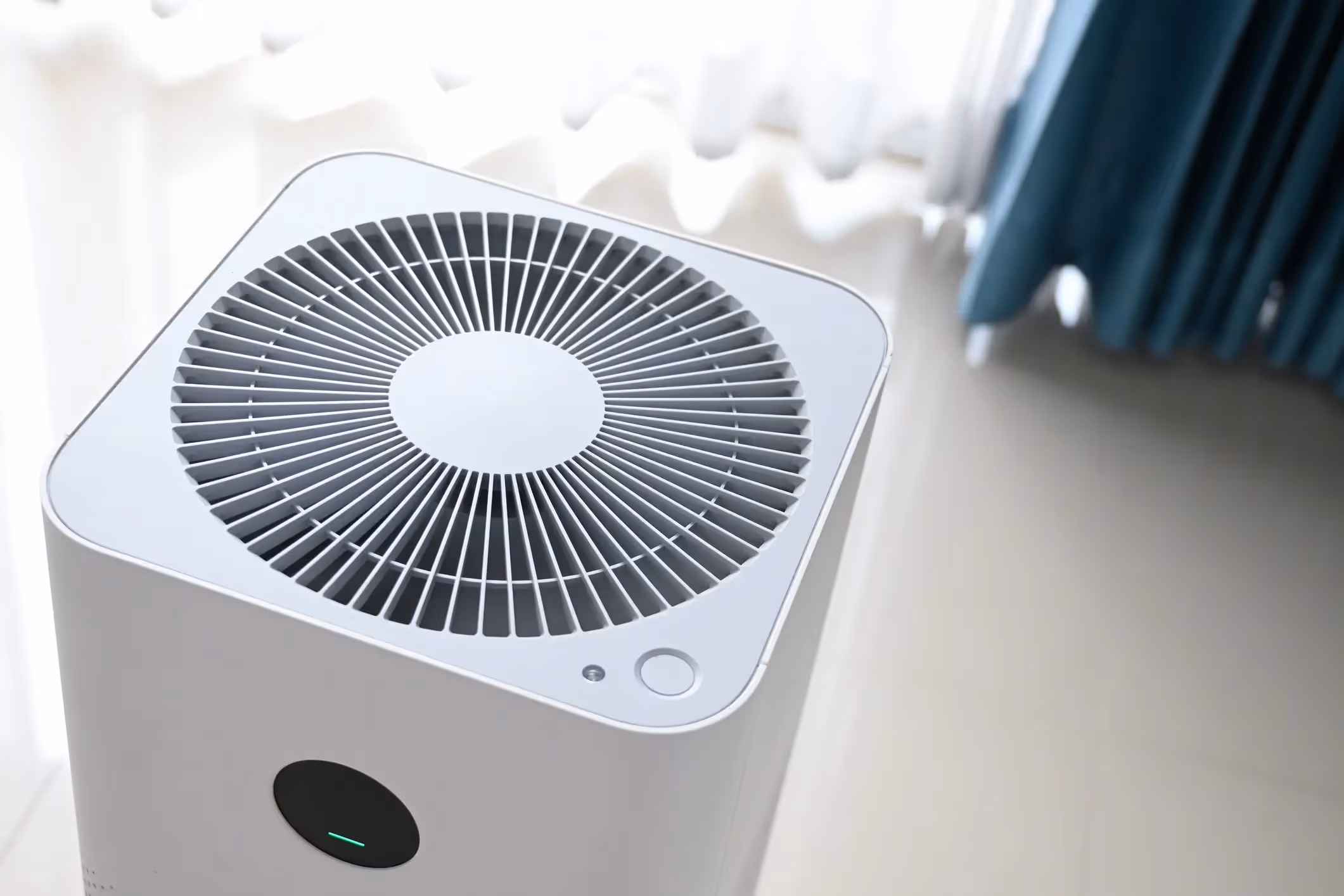 A top-down view of a modern, white, square air purifier sitting on a shiny, light-colored floor. The top of the purifier has a large circular grate. A small, green light on the control panel indicates that it is on. A sheer white curtain and a light blue curtain are visible in the background.
