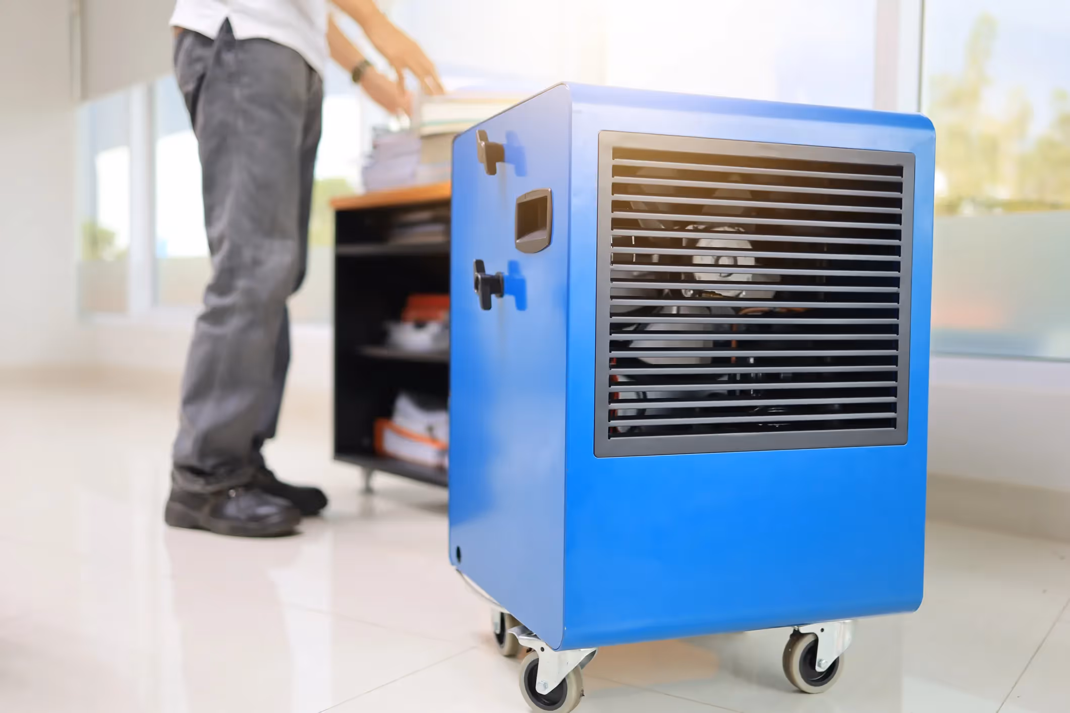 A large, bright blue industrial-style dehumidifier on wheels is positioned on a tiled floor. A person wearing a white shirt and gray jeans is in the background, reaching for something on a low, dark cabinet. The setting appears to be an office or commercial space with large windows and a bright, well-lit interior.