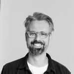 Úsměvavý muž s brýlemi, vousy a krátkými vlasy na světlém pozadí.