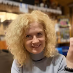 Usměvavá žena s kudrnatými blond vlasy sedí v interiéru s rozostřeným pozadím.