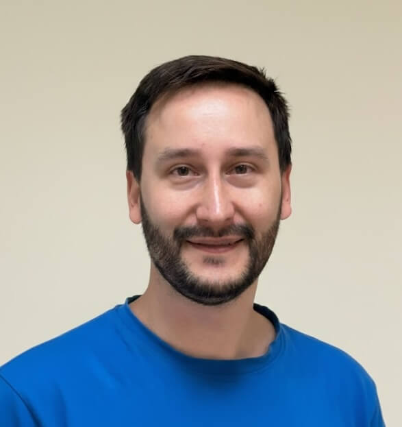 Muž s krátkými tmavými vlasy a vousy v modrém tričku se usmívá před světle béžovým pozadím.