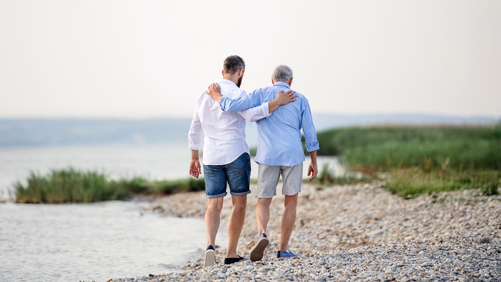 Zwei Männer, die Arm in Arm am Kiesstrand entlang spazieren, mit Blick auf das Wasser und Gras am Ufer.